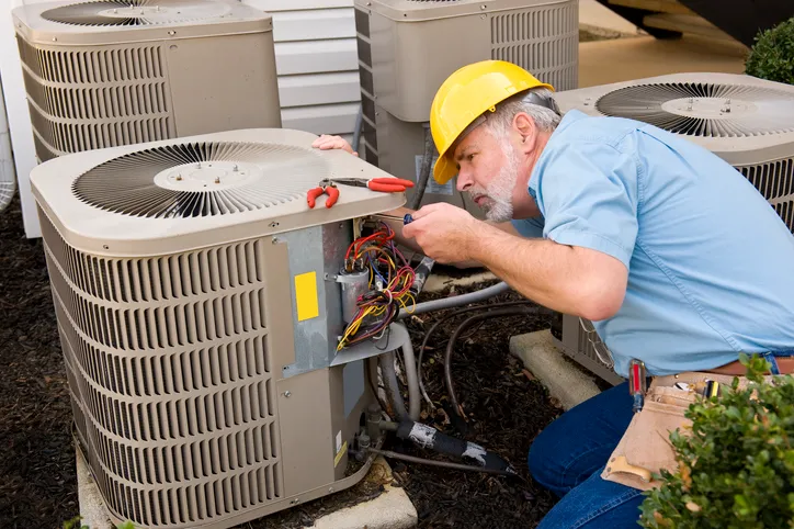AC Installation and Replacement in Bucoda, WA 1 A technician wearing a hard hat, working on the wiring of an outdoor air conditioner unit.