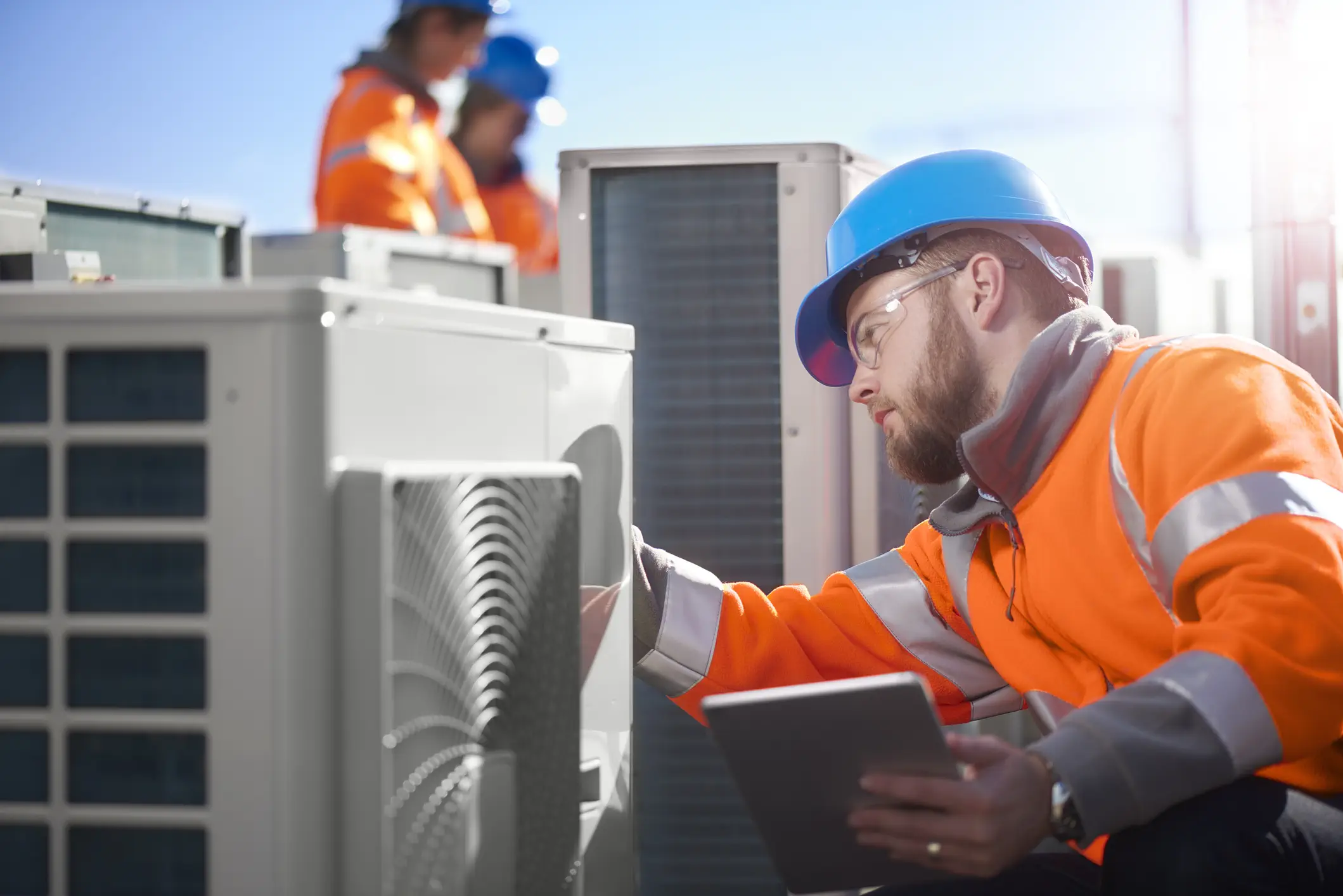 Expert HVAC Installation And Replacement In Bonney Lake, WA 1 A focused male HVAC technician, wearing a bright orange jacket and a blue hard hat, inspects an outdoor air conditioning unit while holding a tablet. In the background, two other workers in similar safety gear are partially visible near more AC units on a sunny rooftop.
