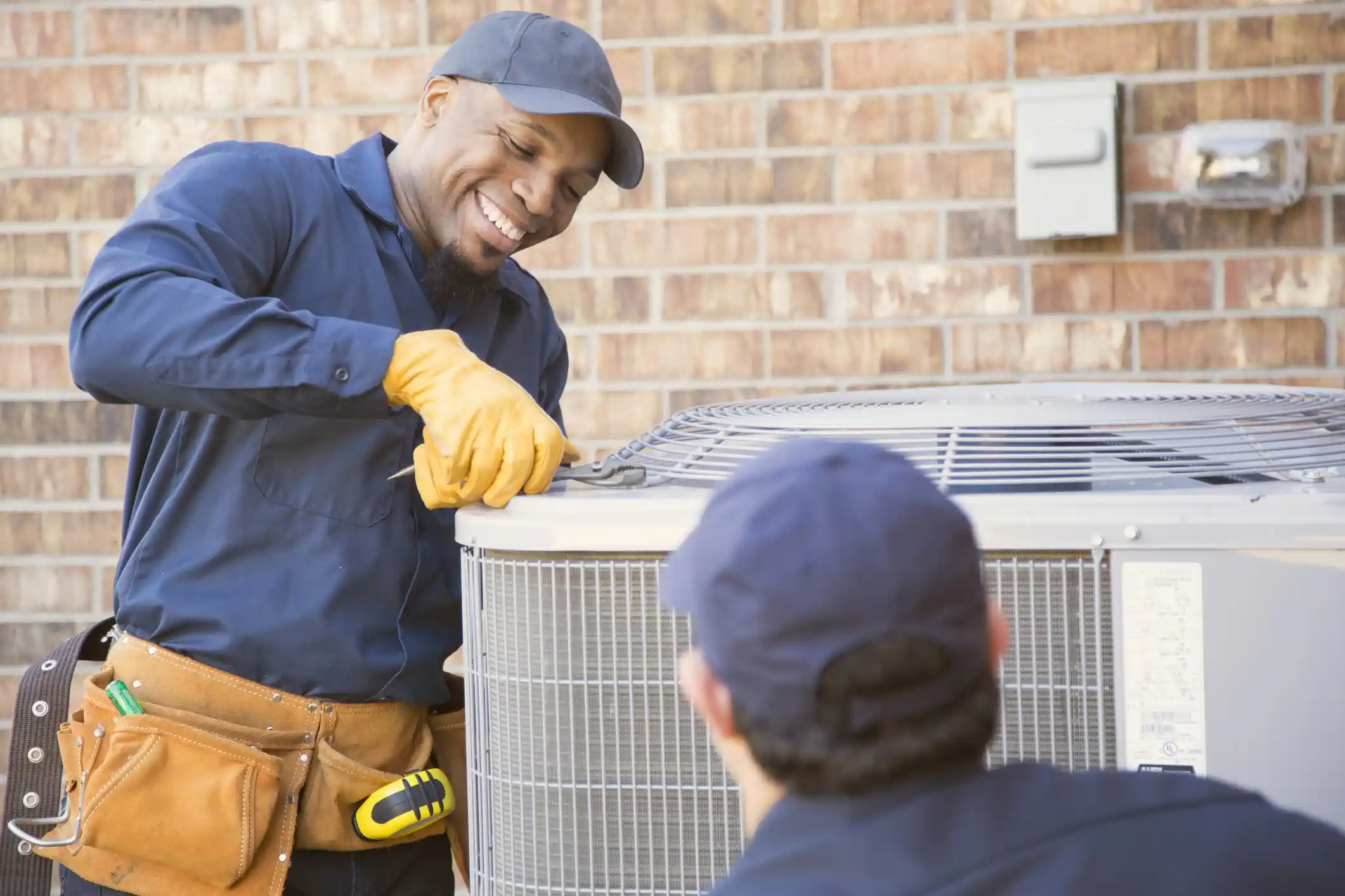 Professional HVAC Installation And Replacement In Bucoda, WA 1 A smiling HVAC technician, wearing a navy blue uniform, cap, tool belt, and yellow safety gloves, uses pliers to work on the top of an outdoor air conditioning unit. Another technician, seen from the back wearing a dark cap, kneels facing the unit. The background features a brick wall.