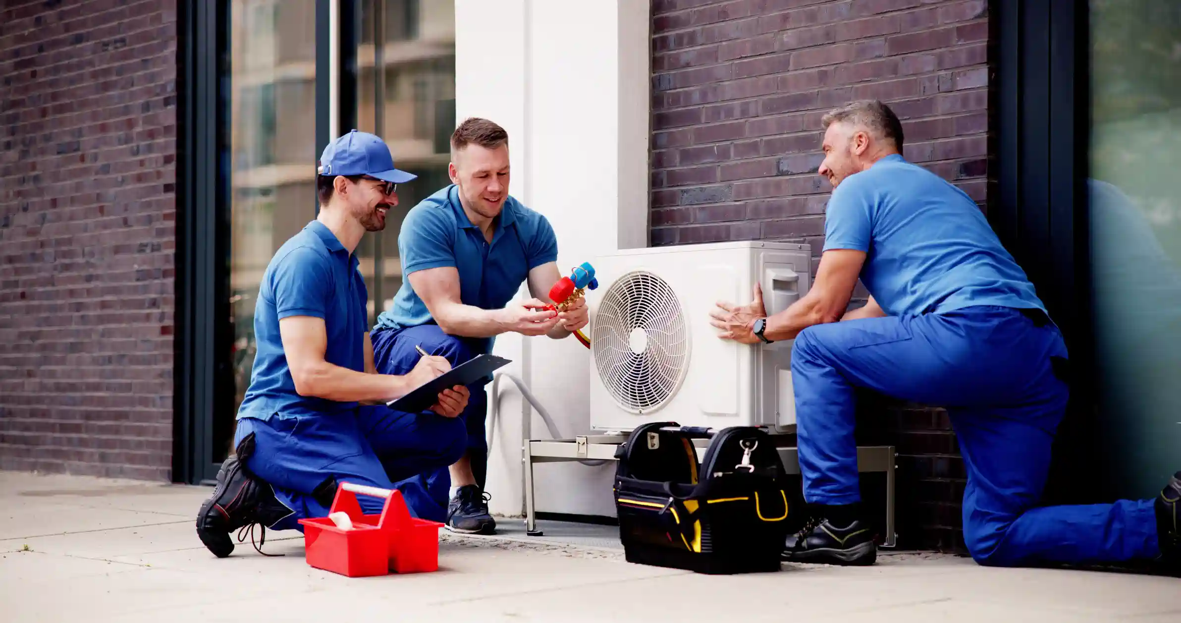 Three smiling HVAC technicians in matching blue uniforms and caps are installing an outdoor air conditioning unit. One man on the right positions the unit, while the man in the center holds refrigerant gauges, and the man on the left holds a clipboard. Two toolboxes are on the ground near the unit.