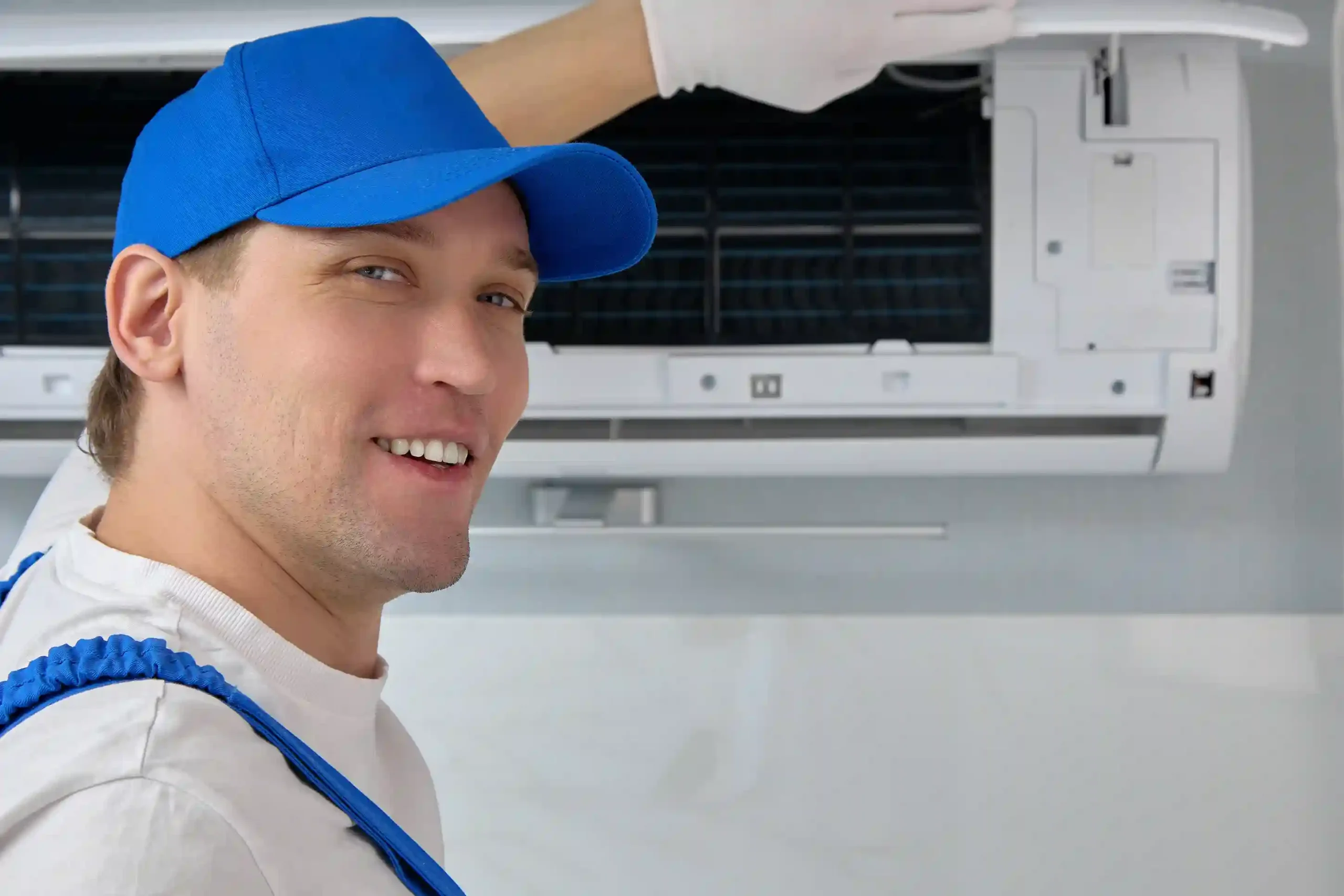 AC Repair in Auburn, WA 3 A happy technician wearing a blue hat and work clothes is standing in front of an open wall-mounted air conditioner.