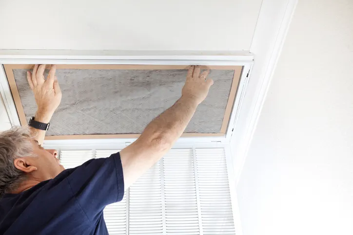 AC Repair in Lakewood, WA 3 A person's hands are replacing a dusty, gray air filter in a ceiling vent.