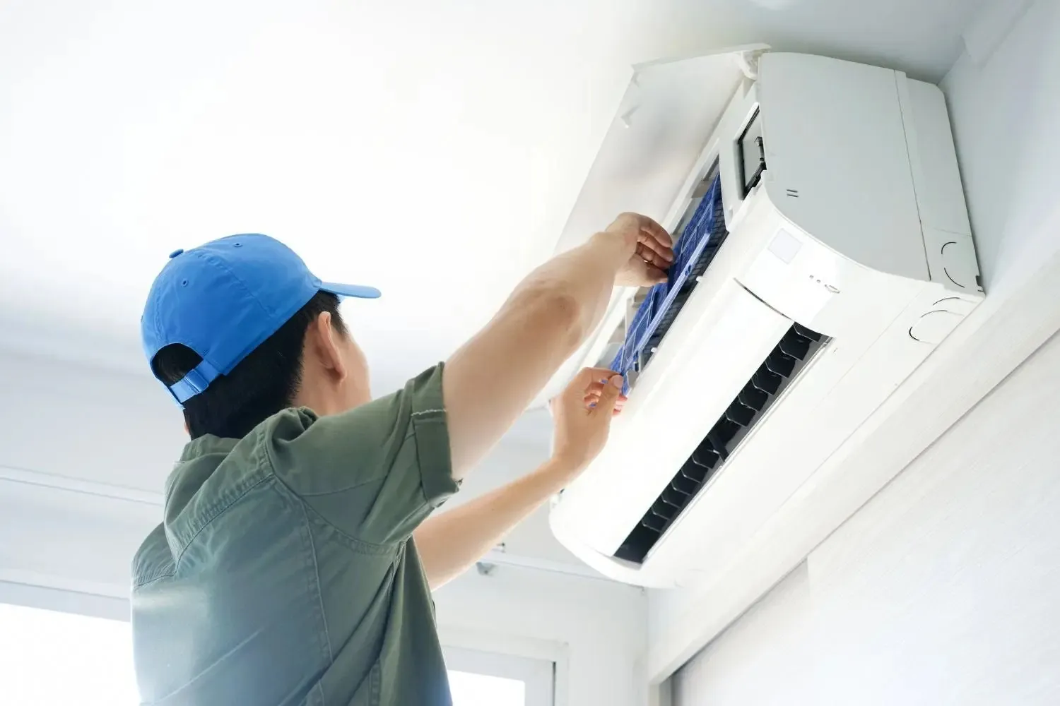 AC Repair Grand in Mound, WA 1 A technician wearing a baseball cap inspects the filter of a white, wall-mounted indoor AC unit.