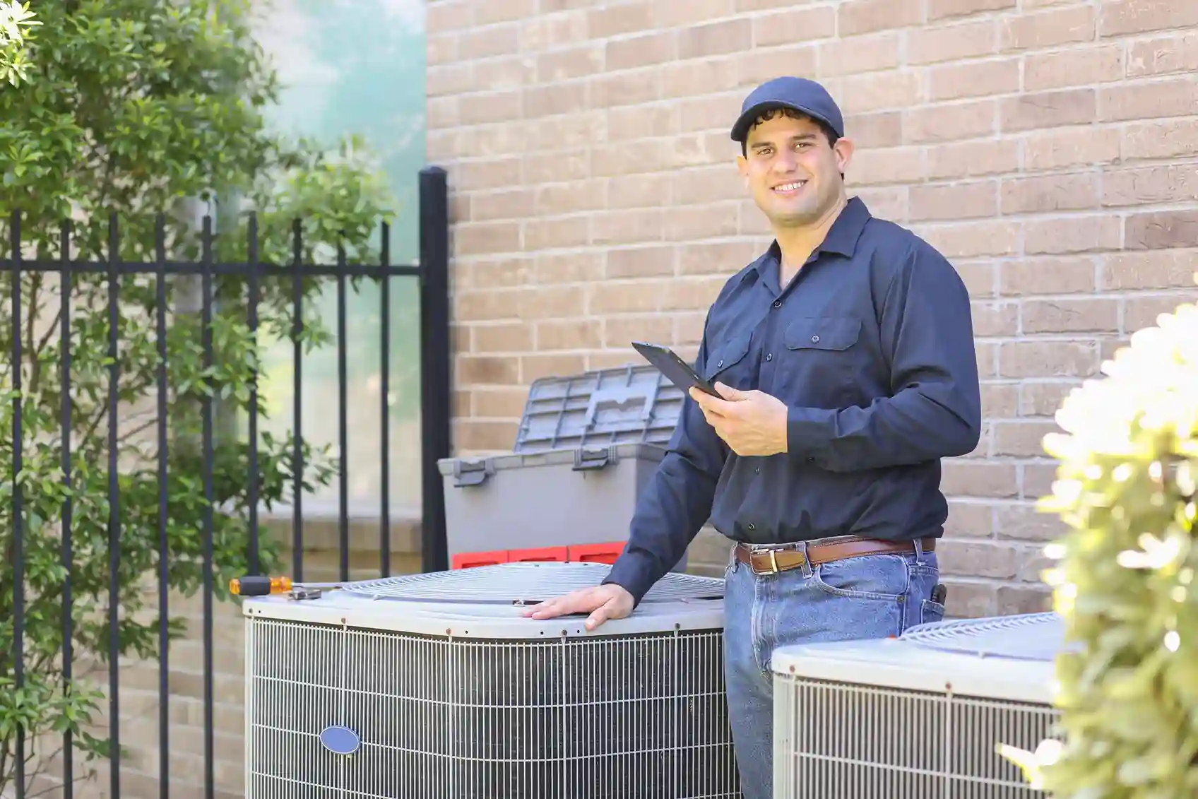 AC Service and Maintenance in Tenino, WA 1 A smiling technician in a blue shirt and cap holding a tablet, standing next to an outdoor air conditioner unit.