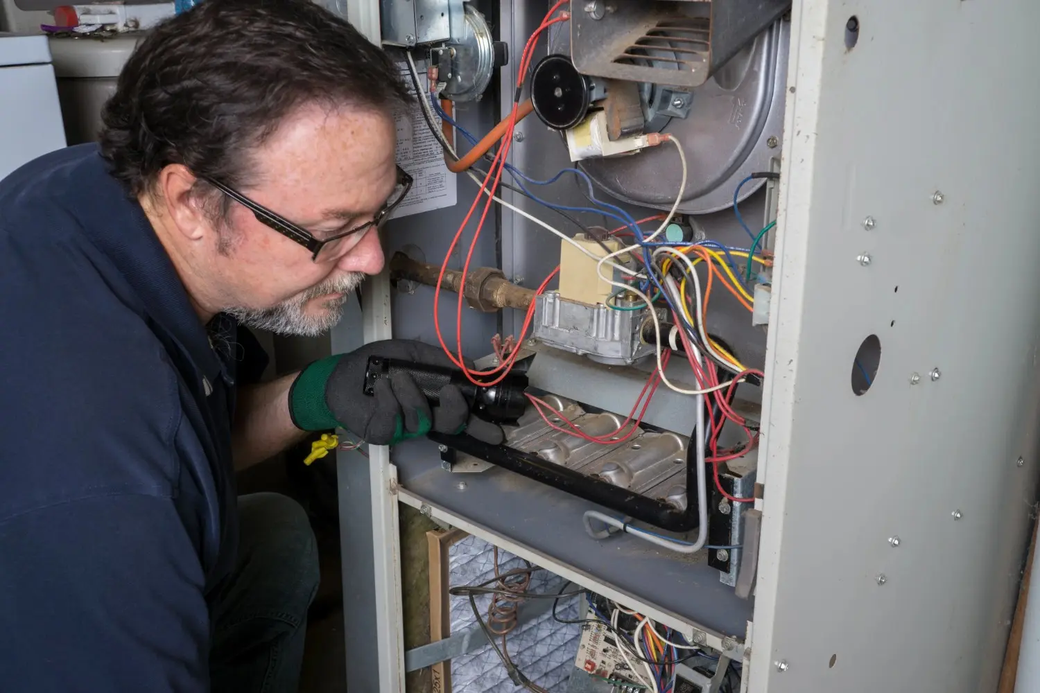 A repairman wearing glasses and a glove inspects the inner workings and wiring of a residential furnace with a flashlight.