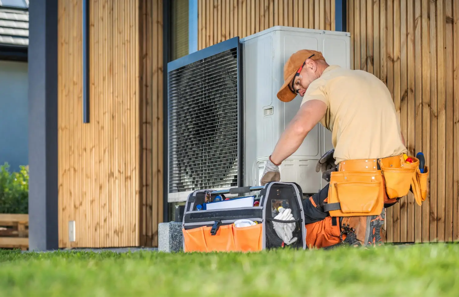 Heating Repair in Tenino, WA 2 A technician in an orange tool belt and cap kneels to perform maintenance on an outdoor heat pump unit next to a modern wood-paneled building.
