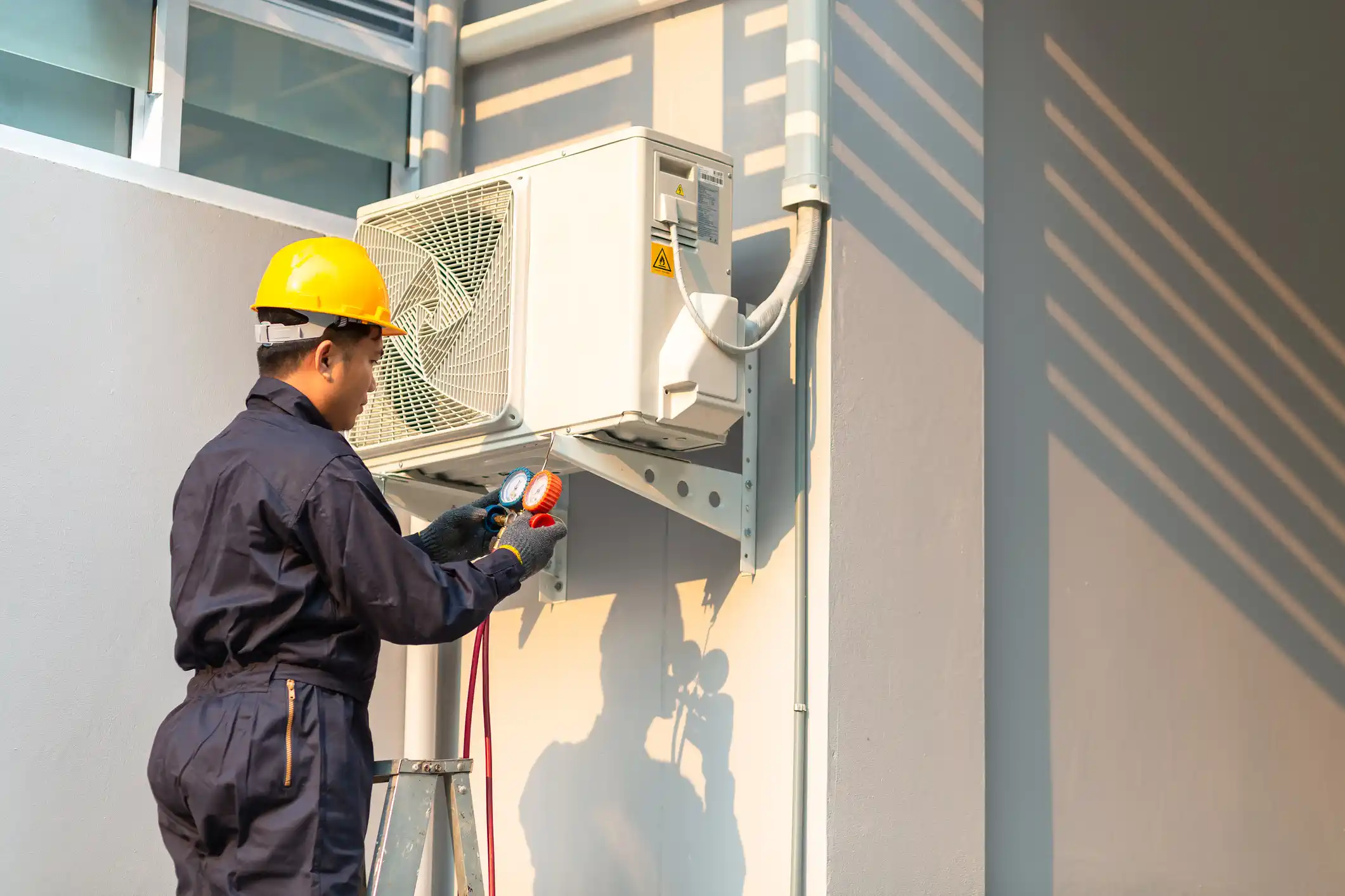 A male technician wearing a dark blue uniform and a yellow hard hat is standing on a ladder and checking an outdoor air conditioning unit mounted on a gray wall. He is holding manifold gauges connected to the unit.