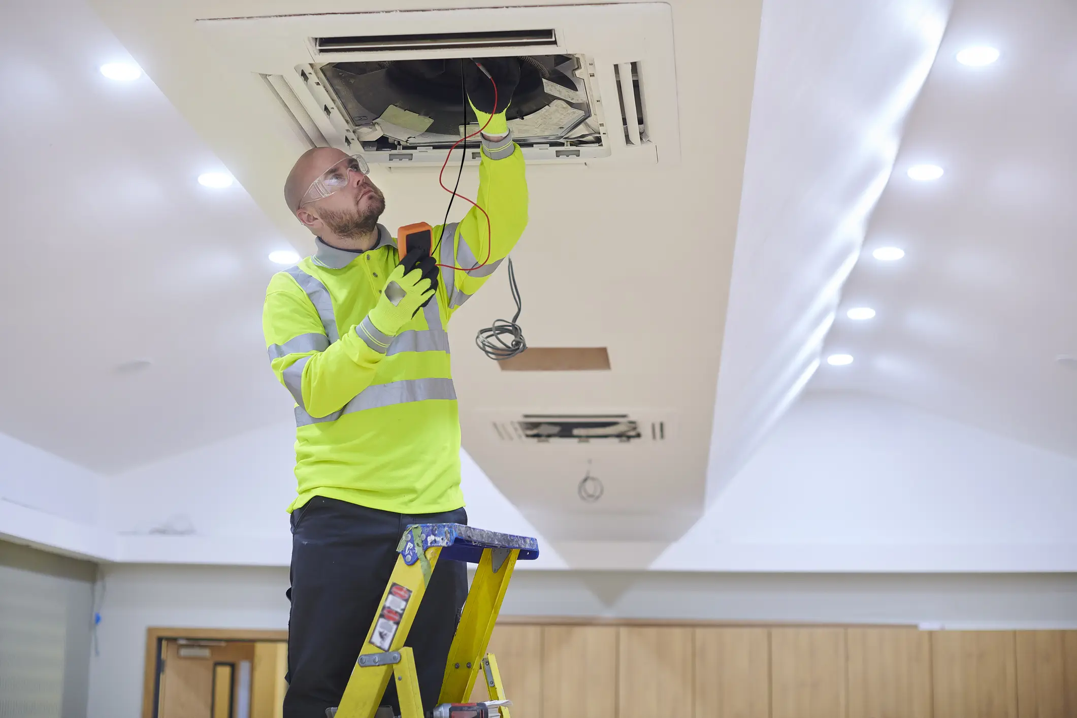 A male technician in a high-visibility yellow-green shirt, black pants, and clear safety glasses is standing on a ladder and working on a ceiling-mounted cassette air conditioning unit. He is holding a digital multimeter in his left hand and touching exposed wires with his right hand.