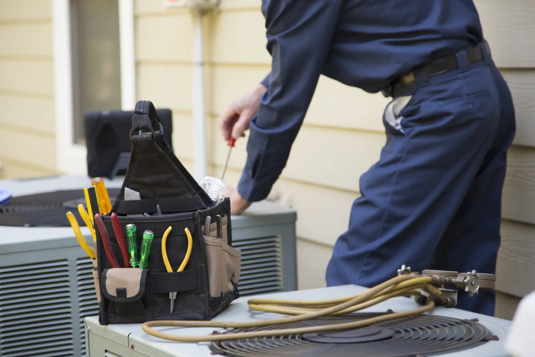 A close-up view of an HVAC technician working on an outdoor air conditioning condenser unit. In the foreground, a tool bag filled with various tools, including pliers and screwdrivers, rests on top of the unit's grille. In the background, a person wearing a dark blue uniform is bent over, using a tool to service or repair the AC unit. There are refrigerant hoses lying next to the tool bag.