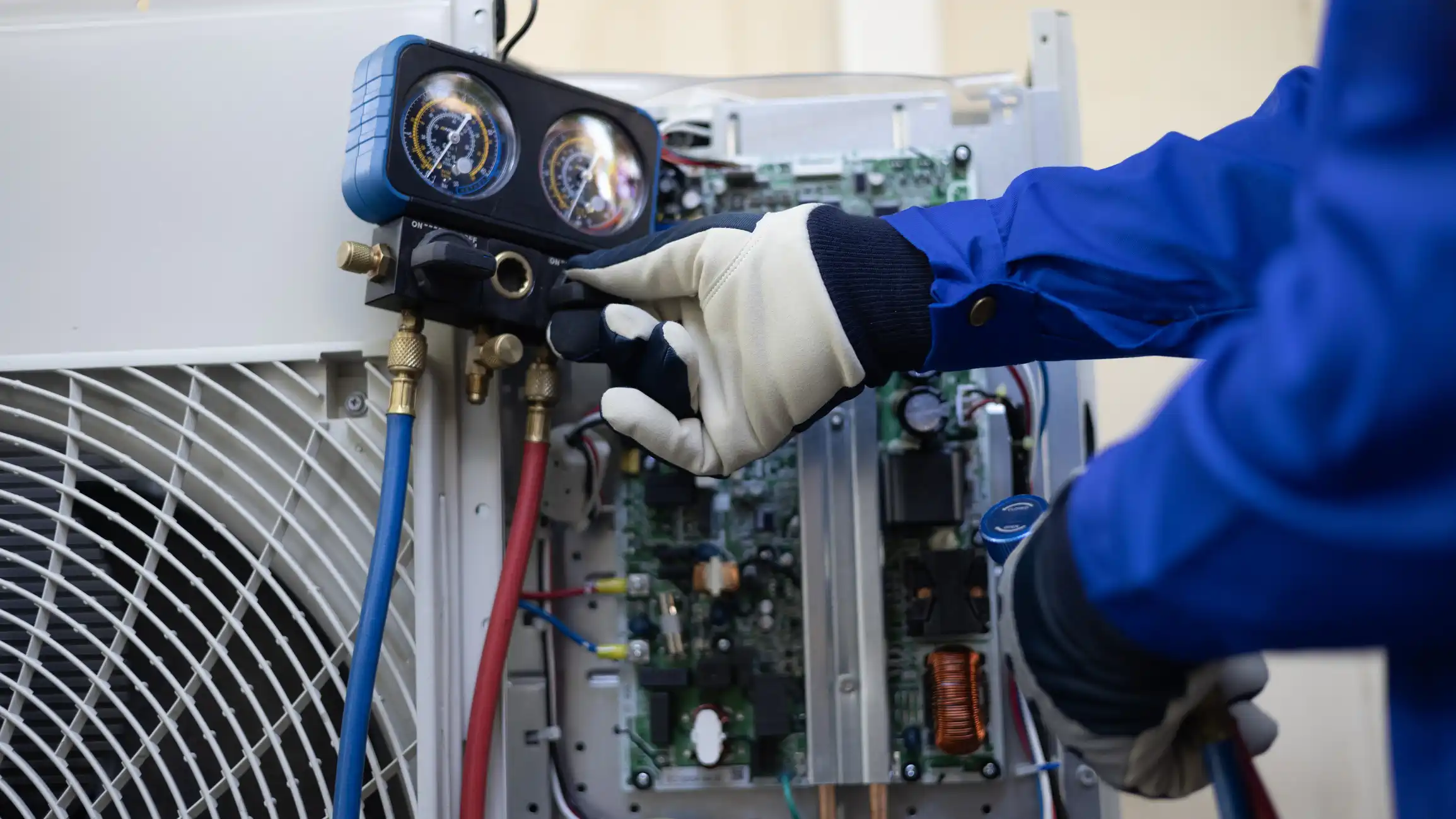 A close-up view of an HVAC technician wearing a white and black glove while adjusting the valves on a refrigerant manifold gauge set. The gauges are connected via blue and red hoses to the service ports of an outdoor air conditioning unit's internal components, which are exposed in the background, showing a circuit board and other electrical parts.