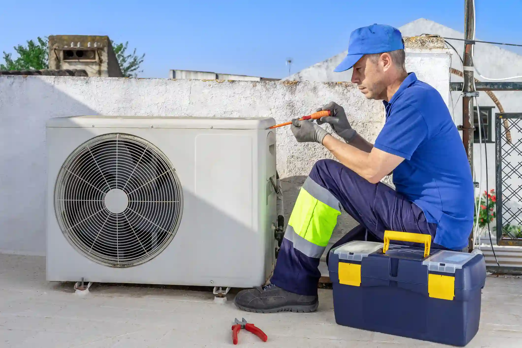 A male HVAC technician wearing a blue uniform, blue baseball cap, safety gloves, and reflective work pants is crouched on a flat, light-colored roof or patio, working on a white outdoor air conditioning unit (a mini-split condenser). He is using a screwdriver to service the unit. A blue and yellow toolbox is placed on the ground next to him, along with a pair of red-handled pliers.