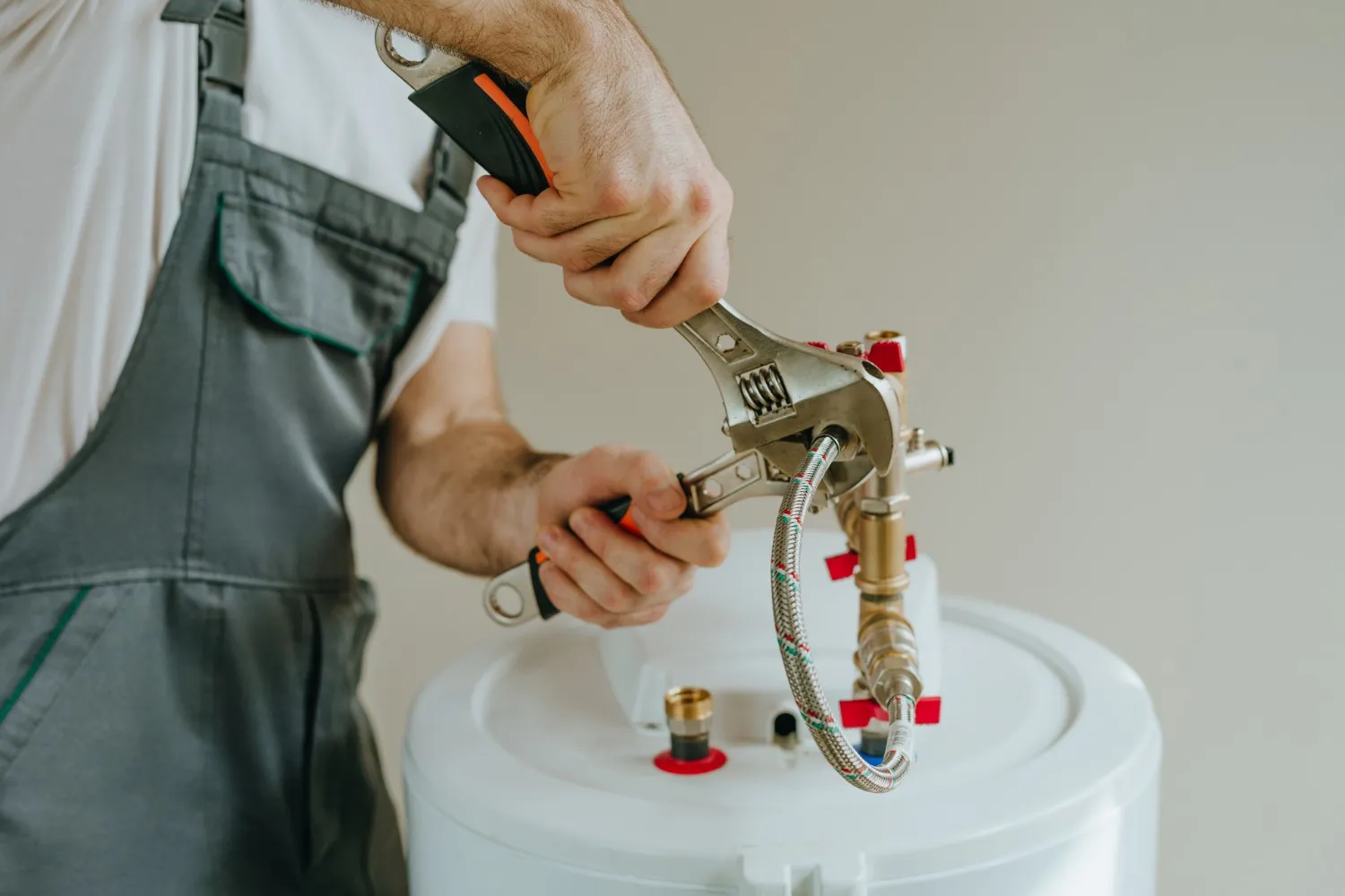 Professional HVAC Installation And Replacement In Enumclaw, WA 3 A close-up shot shows a worker, wearing grey overalls and a white shirt, using a large adjustable wrench to tighten fittings and a braided metal hose connecting to the top of a white water heater or boiler unit.