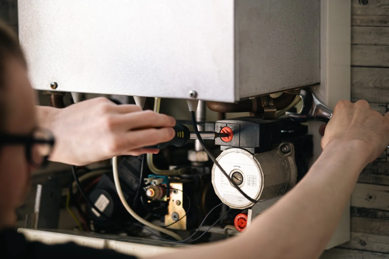 A close-up over-the-shoulder view shows a technician with glasses servicing the internal components of a gas boiler or heating unit. The person is using a screwdriver to adjust a part of the internal mechanism, which includes wiring, valves, and a circulation pump.