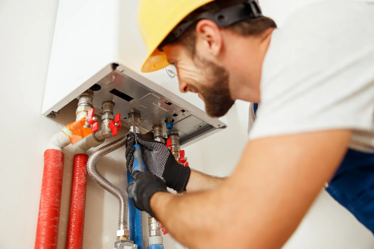A smiling male technician, wearing a white shirt, blue overalls, a yellow hard hat, and black gloves, is using a wrench to service the piping connections on the underside of a wall-mounted white boiler or water heater. Red-capped valves and thick red and blue insulated pipes are visible below the unit.