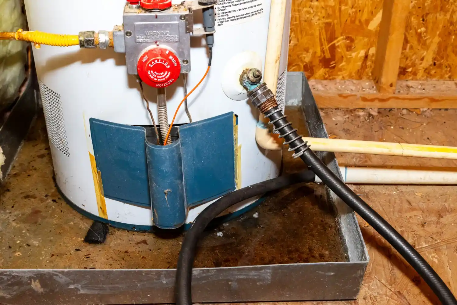 A close-up image shows the lower portion of a white gas water heater sitting in a metal drain pan. A black hose is connected to the water heater's drain valve, with a small puddle of water visible in the pan. The gas control valve is prominently featured with an orange wire connected to the thermocouple.