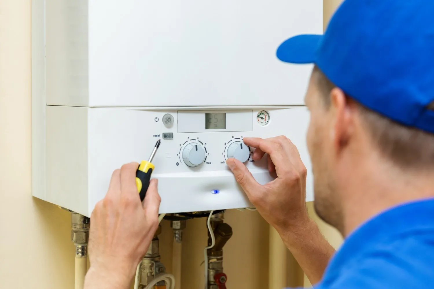 A close-up image shows a technician, wearing a blue shirt and cap, adjusting the controls on the front panel of a white wall-mounted gas boiler or heating unit. He is turning one of the temperature knobs with his right hand and holding a screwdriver near the reset button with his left hand.