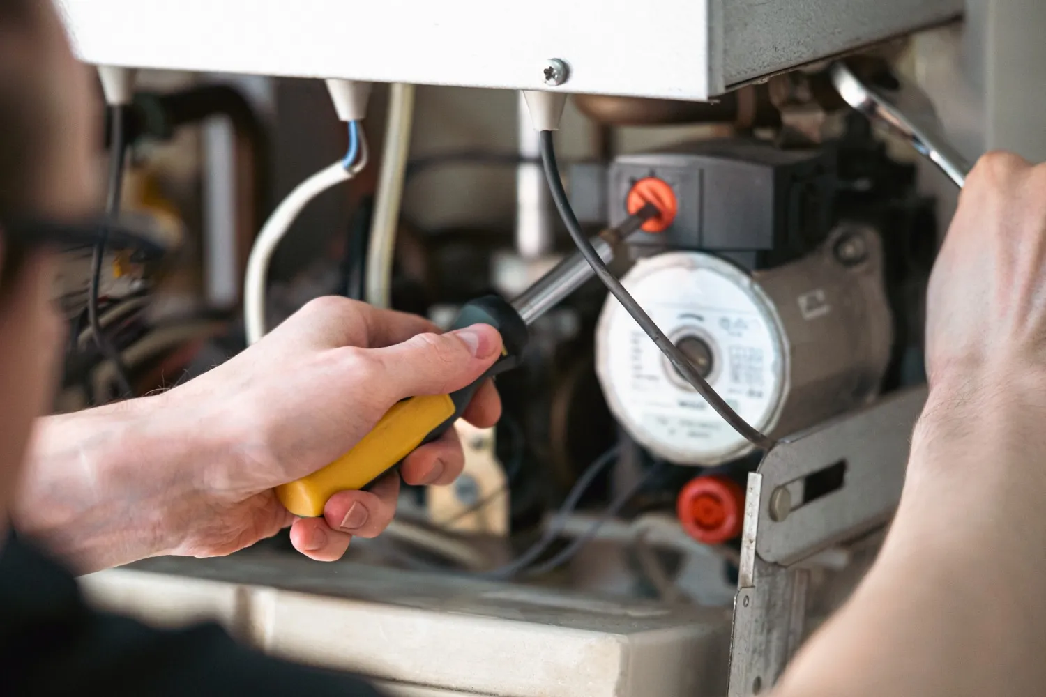 A close-up, over-the-shoulder view shows a technician with glasses servicing the internal components of a gas boiler or heating unit. The person is holding a yellow and black screwdriver in their right hand, adjusting a part, while using a wrench with their left hand to work on another component near the circulation pump.