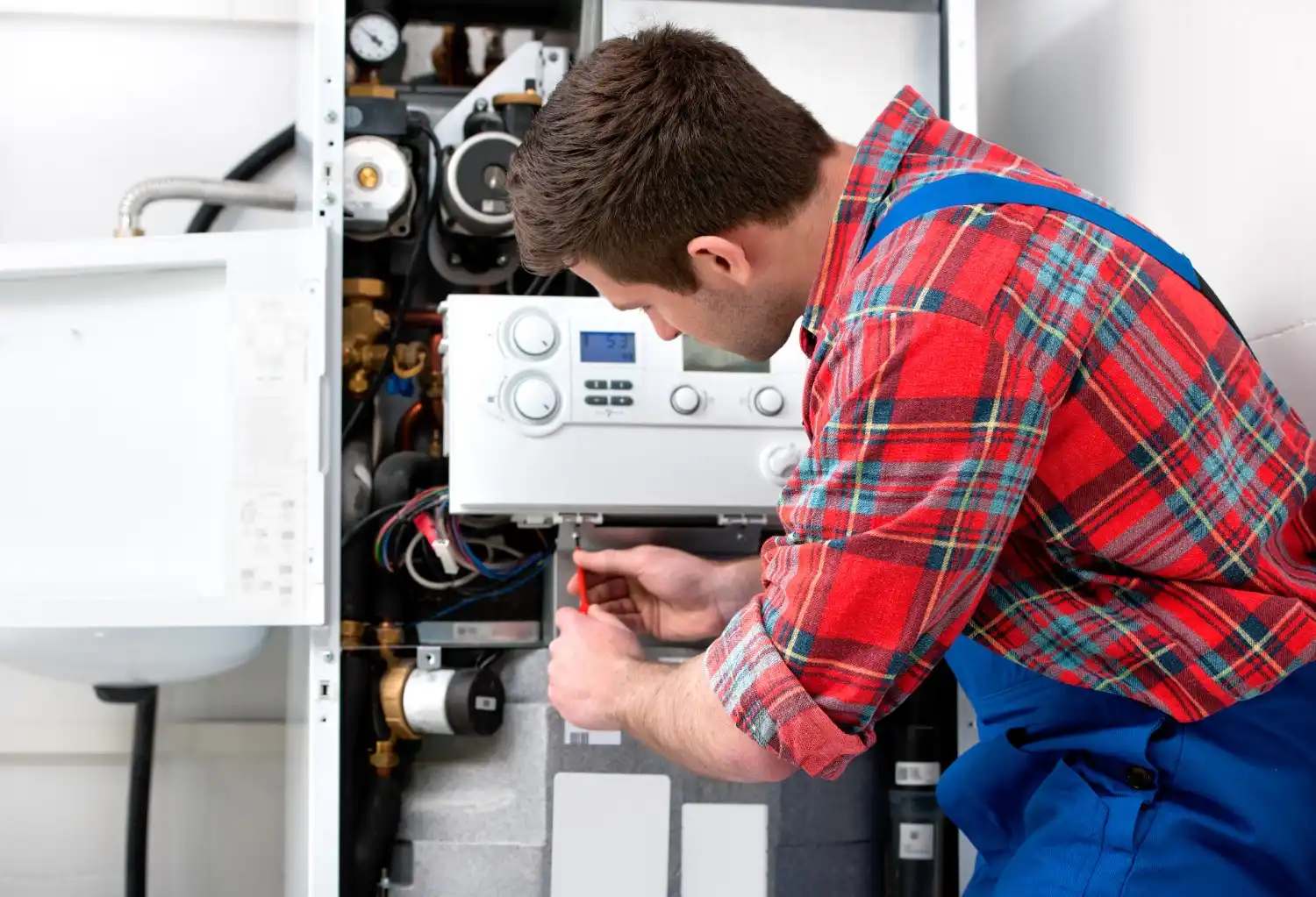 A male technician, wearing a red plaid shirt and blue overalls, is focused on repairing the internal components of a wall-mounted boiler. He is using a small screwdriver to adjust a wire or component near the control panel, which features a digital display. Various pipes and gauges are visible around the unit.