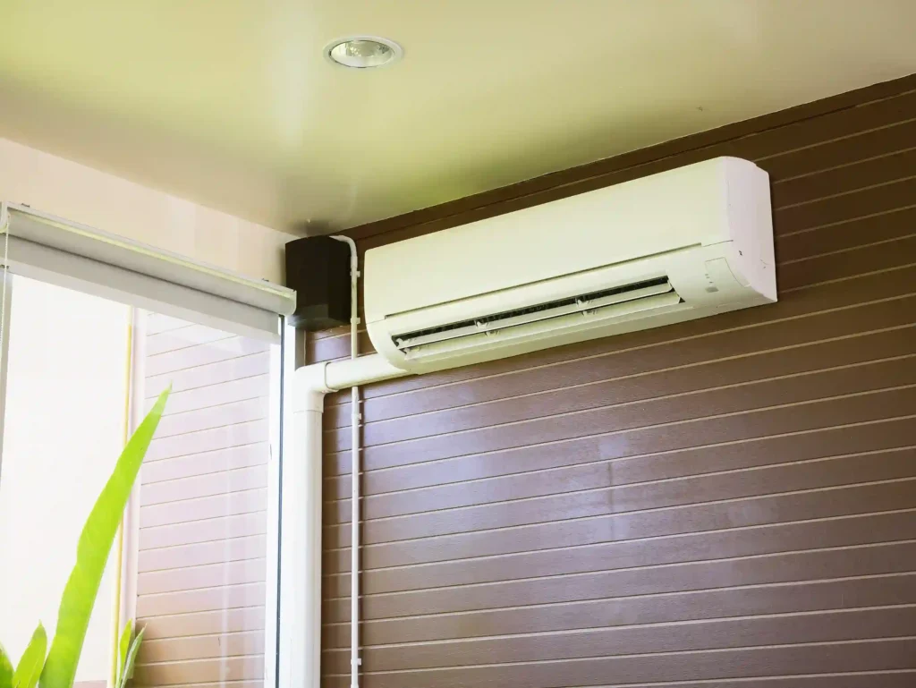 A white ductless mini-split indoor air handler unit is mounted high on a wall with brown horizontal paneling. White piping and a cable conduit run down from the unit. The room has a light-colored ceiling with a recessed light and a large window with white blinds is visible to the left.