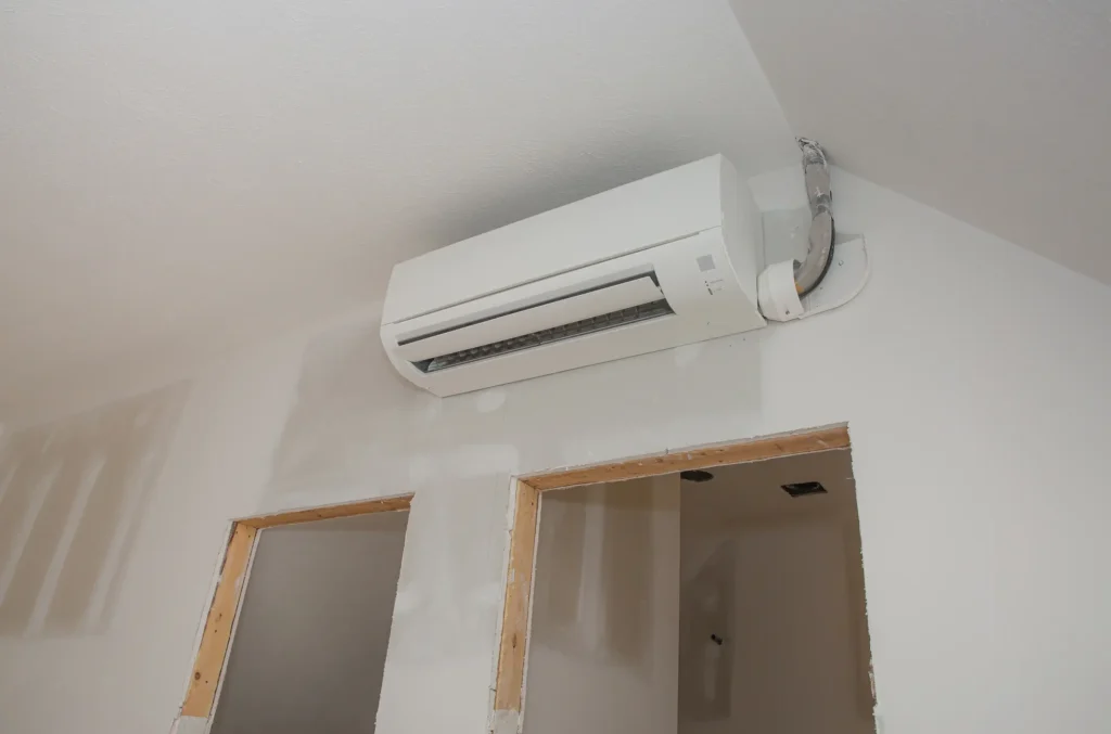 A white ductless mini-split indoor air handler unit is mounted high on a light gray wall in a room under construction, with unpainted drywall visible. The unit is placed above two adjacent, unfinished doorways with exposed wood framing. The refrigerant lines and wiring are visible entering the unit on the right.