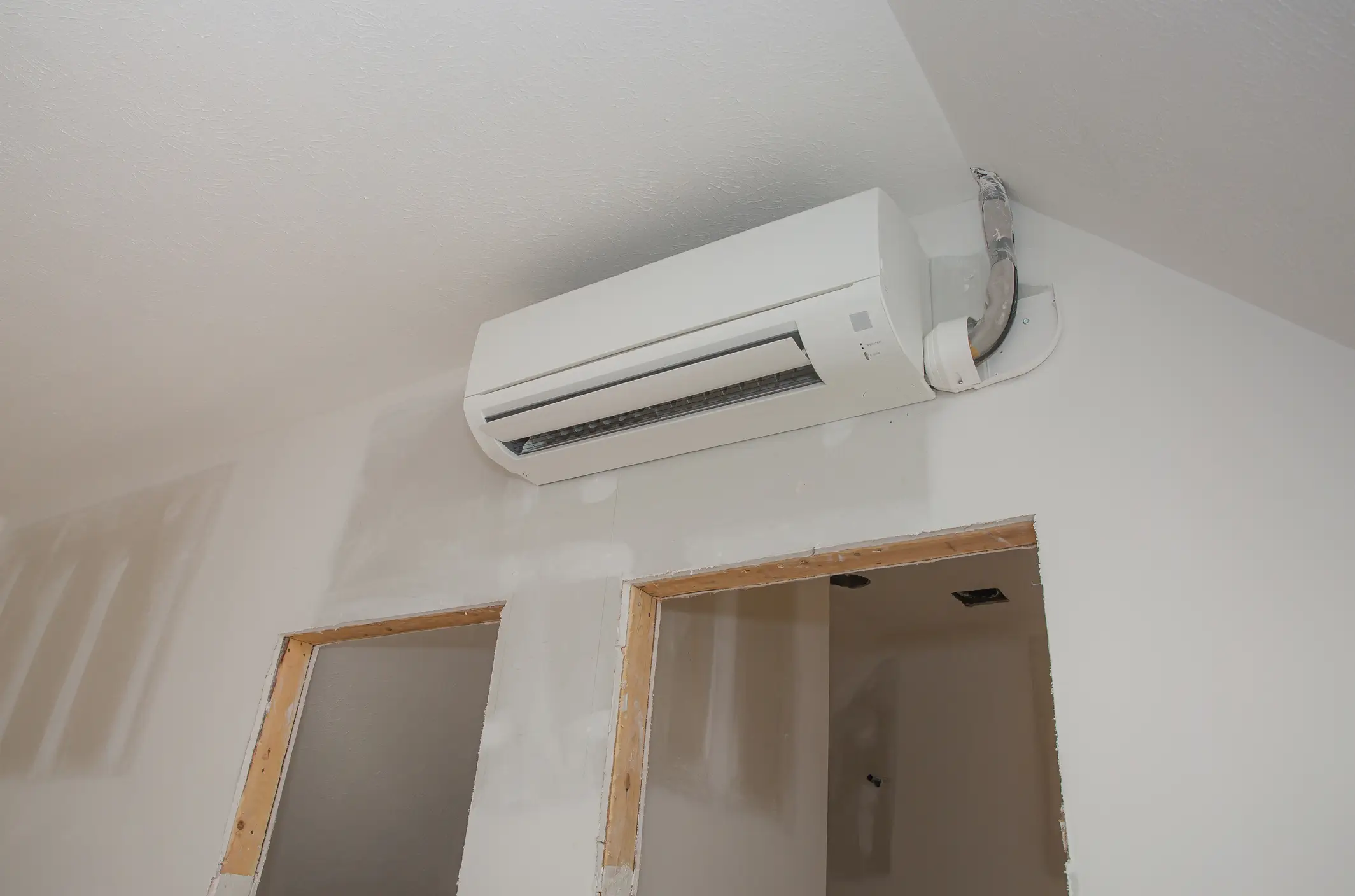 A white ductless mini-split indoor air handler unit is mounted high on a light gray wall in a room under construction, with unpainted drywall visible. The unit is placed above two adjacent, unfinished doorways with exposed wood framing. The refrigerant lines and wiring are visible entering the unit on the right.