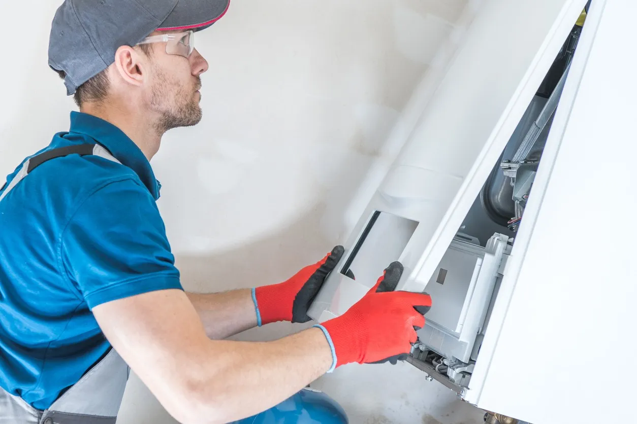 A male technician, wearing a blue shirt, gray pants, a dark cap, safety glasses, and red and black gloves, is opening the front panel of a white wall-mounted boiler or furnace unit. The technician is focused on accessing the internal components for repair or maintenance.