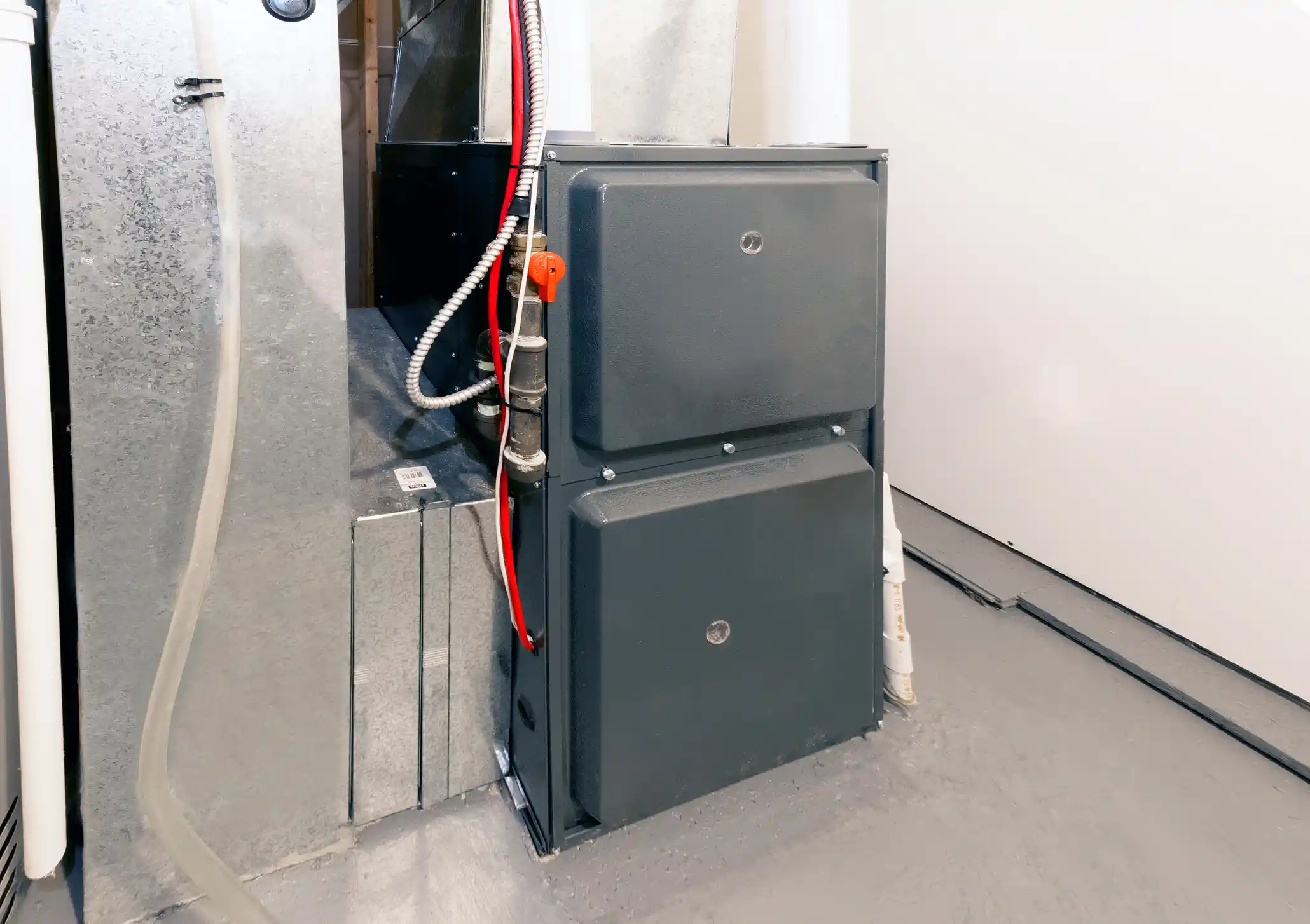 A residential gas furnace, featuring two dark gray access panels, stands next to a large metal air duct. A condensate line and a gas line with a red shut-off valve are visible running along the side of the unit. The furnace is located in a utility space with gray painted floors and white walls.