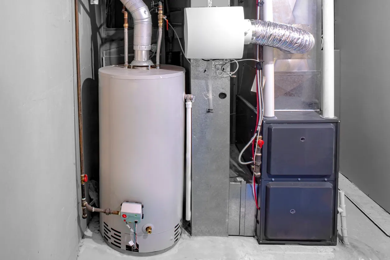 A residential utility corner contains a hot water heater and a furnace. The tall, light gray water heater is on the left, connected to copper piping. To the right is a dark gray furnace with two access panels, connected to large metal and flexible foil air ducts. The walls and floor of the utility space are painted a solid gray color.