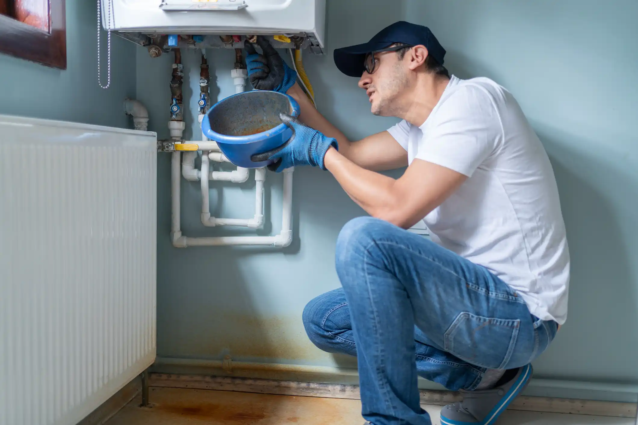 A male technician in a white shirt, jeans, dark cap, and blue work gloves, kneels to repair a wall-mounted white boiler or water heater. He is holding a blue bowl underneath the piping connections to catch dripping water, while simultaneously working on the pipes. A white radiator is visible to the left.