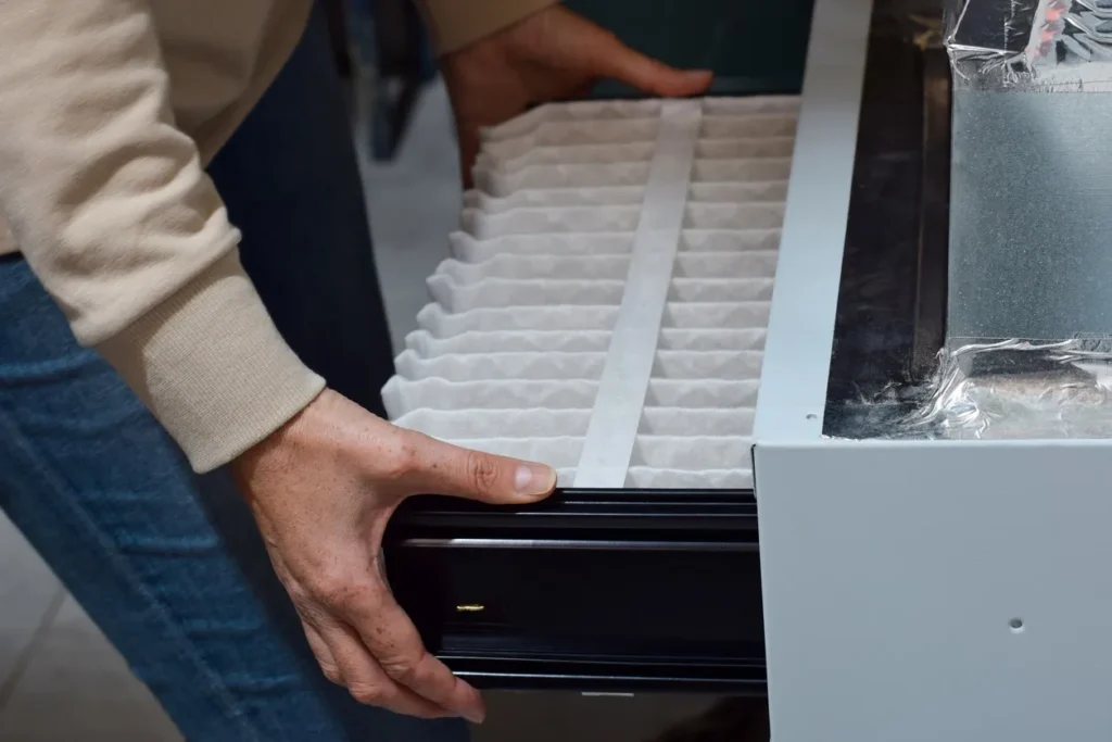 A person wearing a beige sweater and blue jeans is inserting a pleated, rectangular white air filter into the slot of a furnace or HVAC unit. The person's hands are holding the filter frame as it slides into the metal casing.