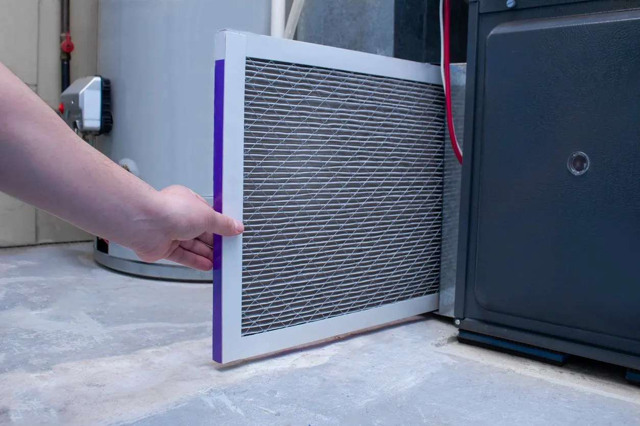 A person's hand is seen inserting a rectangular HVAC air filter, which has a purple border and wire mesh over the filtering material, into the lower slot of a dark gray furnace. A light gray water heater is visible in the background, all resting on a concrete floor.