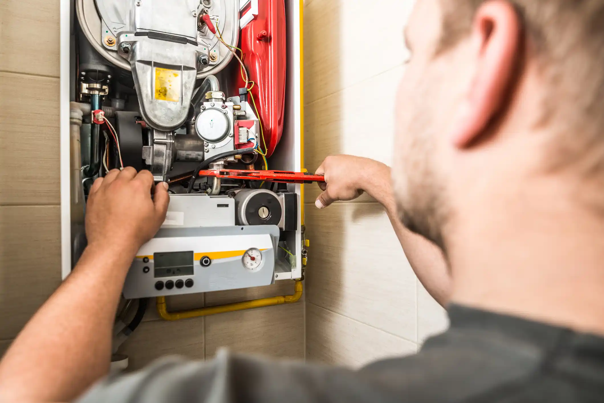 An over-the-shoulder close-up shows a male technician servicing the internal components of a wall-mounted boiler or furnace. He is using a red and black tool, possibly a wrench or pliers, to adjust a part near the heat exchanger and controls. The inside of the unit features various wires, gauges, and mechanical parts.