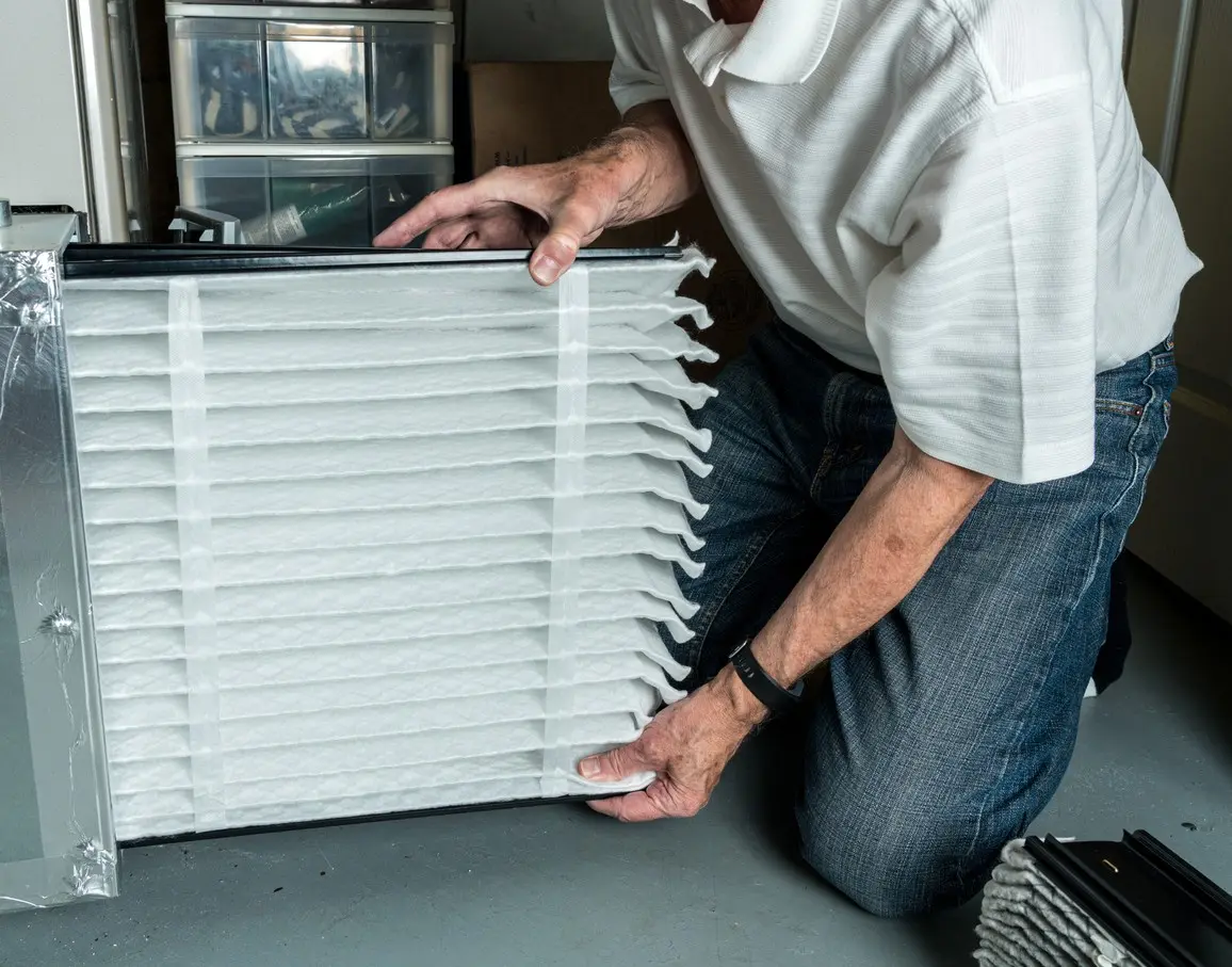 A person, wearing a white polo shirt and jeans, is kneeling and using both hands to insert a large, thick, pleated white air filter into an HVAC air handler unit. The filter is partially inserted into a metal frame, and plastic storage drawers are visible in the background.