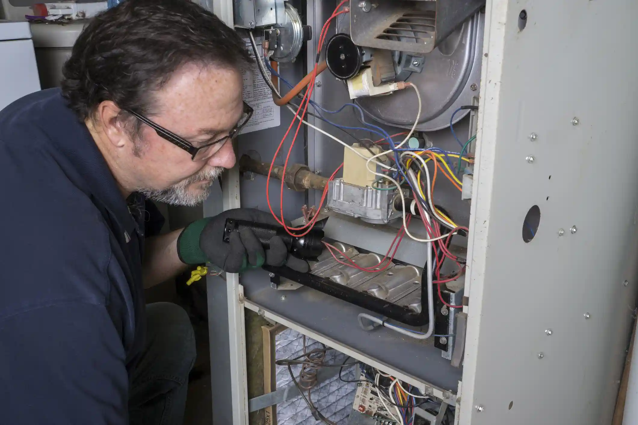 A middle-aged male technician with glasses and a beard is inspecting the open combustion chamber of a furnace. He is wearing a dark blue shirt and green and black gloves, holding a small flashlight to illuminate the burners and heat exchanger components, which are surrounded by various colored wires.