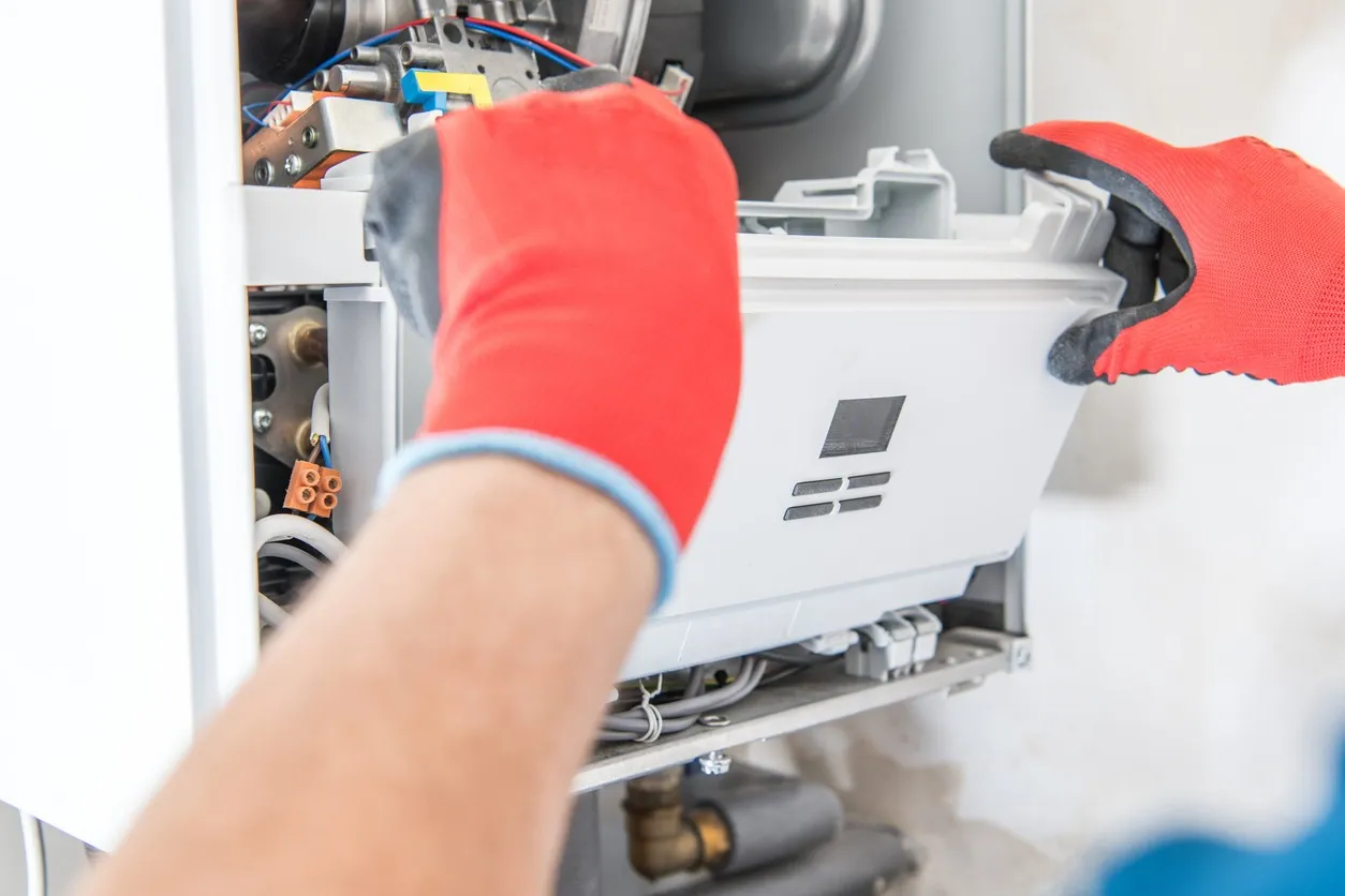 A close-up view shows a technician, wearing red and gray gloves, carefully removing a white plastic component from the internal cavity of a wall-mounted boiler or furnace unit. Various wires and metal parts are visible inside the open unit.