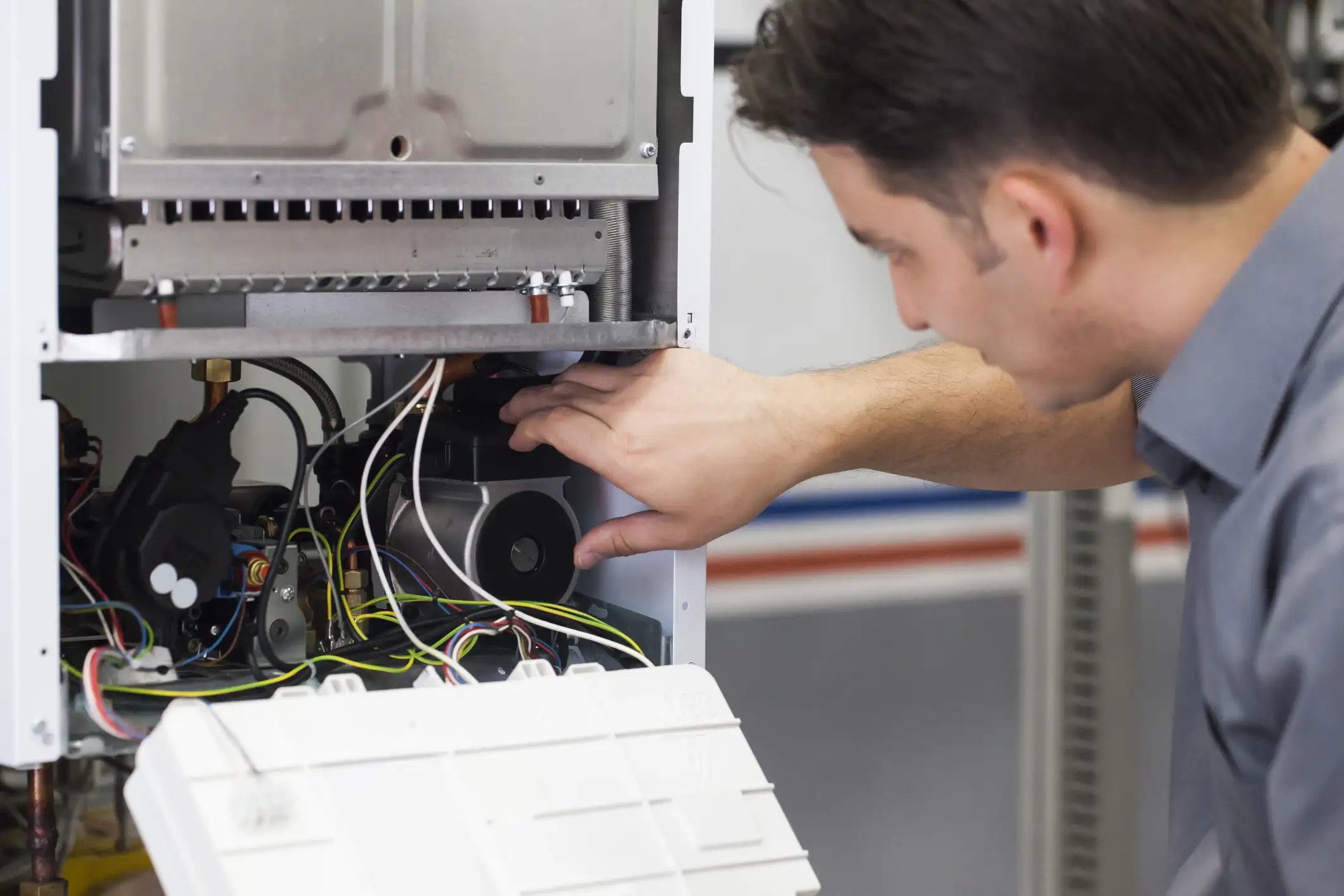 A male technician in a gray shirt is focused on inspecting the internal components of a white wall-mounted boiler or furnace, which has its front panel opened. He is reaching inside to touch a component near the circulation pump, surrounded by various electrical wires.