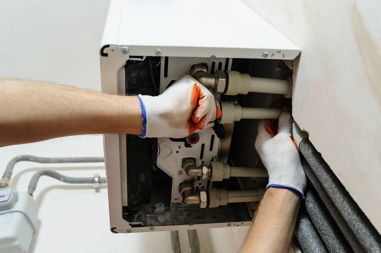 A close-up image shows a technician, wearing white and orange work gloves, connecting white plastic PEX-style piping with brass fittings to the underside of a wall-mounted white water heater or furnace unit. Insulated gray piping is visible entering the wall on the right, and a gas meter is partially visible to the left.
