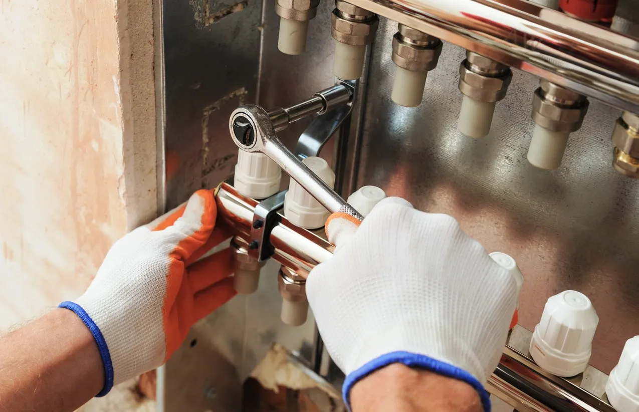 A close-up image shows a worker, wearing white and orange gloves, using a ratcheting wrench to tighten a fitting on a shiny chrome manifold assembly. The manifold connects multiple white plastic pipes, likely for a radiant floor heating system, recessed into a wall space.