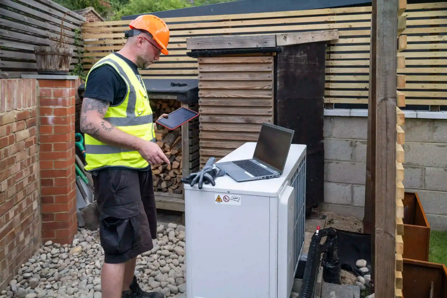 A male HVAC technician wearing a high-visibility vest, dark shorts, and an orange hard hat is standing next to an outdoor heat pump unit. He is holding a tablet in his left hand and is working near a laptop placed on top of the heat pump. A pair of gloves rests on the laptop. The unit is located in a backyard with a brick wall and a wooden fence/shed structure.