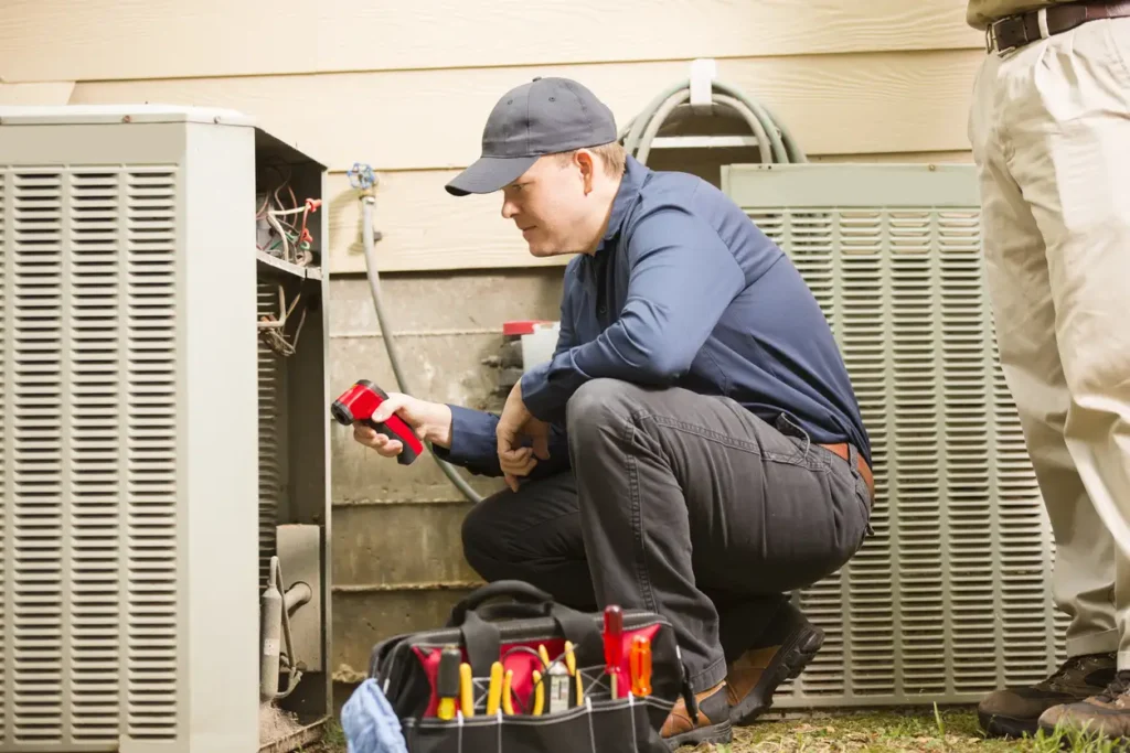 A male technician, wearing a dark blue shirt, black pants, and a dark cap, is crouched down inspecting the open side of an outdoor heat pump or air conditioning unit. He is holding a red testing device. Another person, seen only from the waist down in beige pants, stands next to him. A black tool bag sits open on the ground in the foreground.