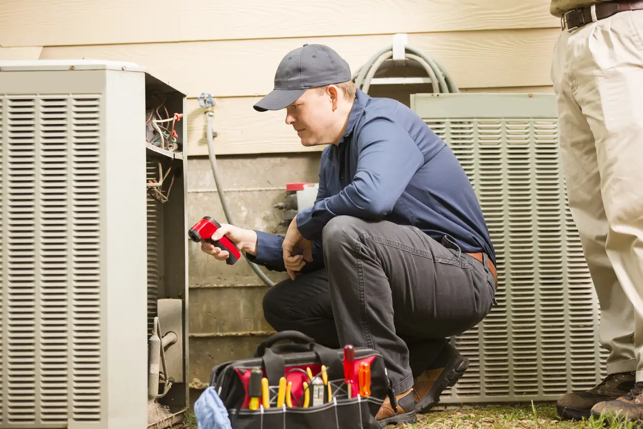 A male technician, wearing a dark blue shirt, black pants, and a dark cap, is crouched down inspecting the open side of an outdoor heat pump or air conditioning unit. He is holding a red testing device. Another person, seen only from the waist down in beige pants, stands next to him. A black tool bag sits open on the ground in the foreground.