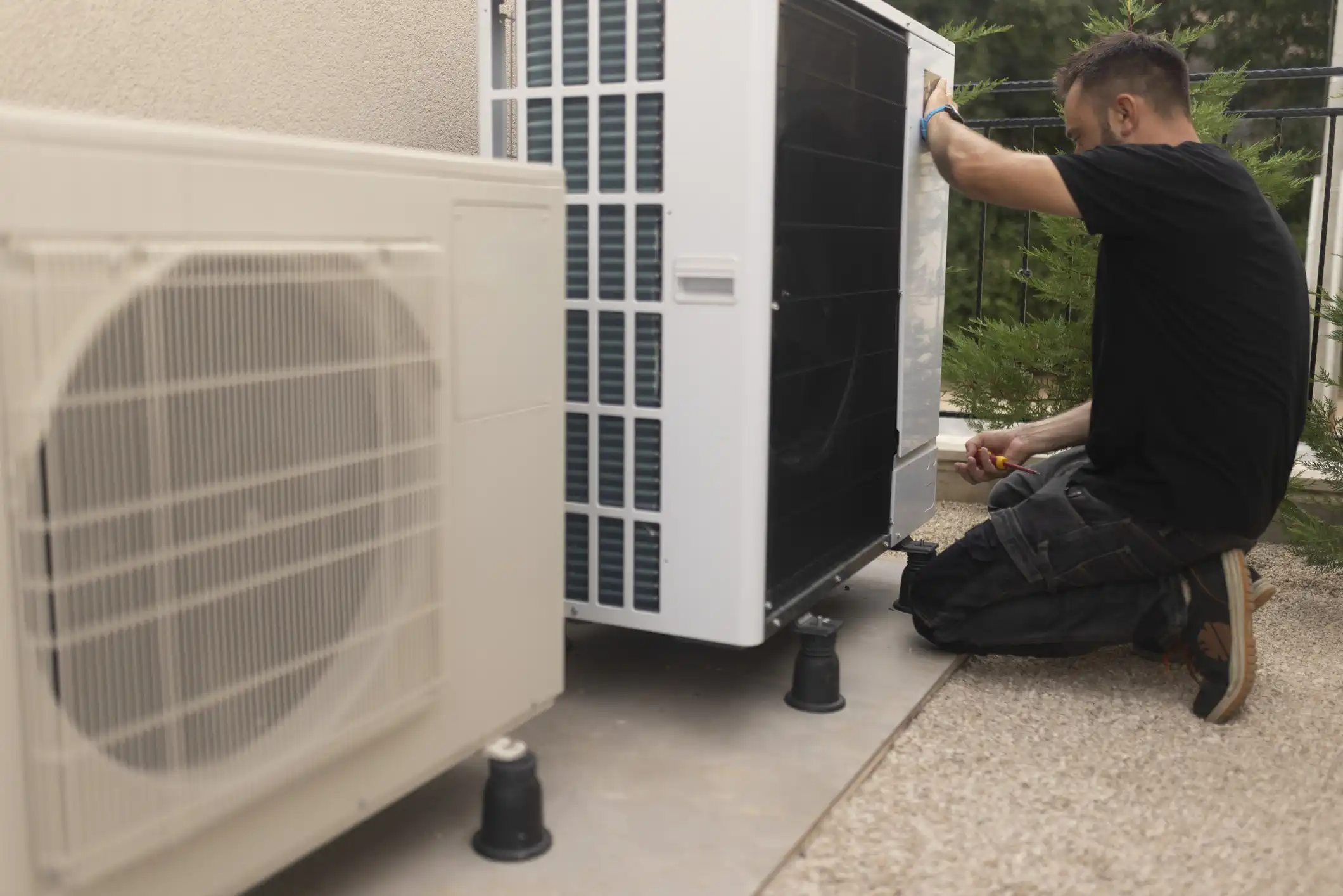 A technician, wearing a black t-shirt and work pants, kneels on a light-colored patio while servicing the outdoor unit of a heat pump system . There are two white heat pump units visible, one larger and one smaller, mounted slightly above the ground on black risers . The technician is focused on the larger, partially black-paneled unit on the right .