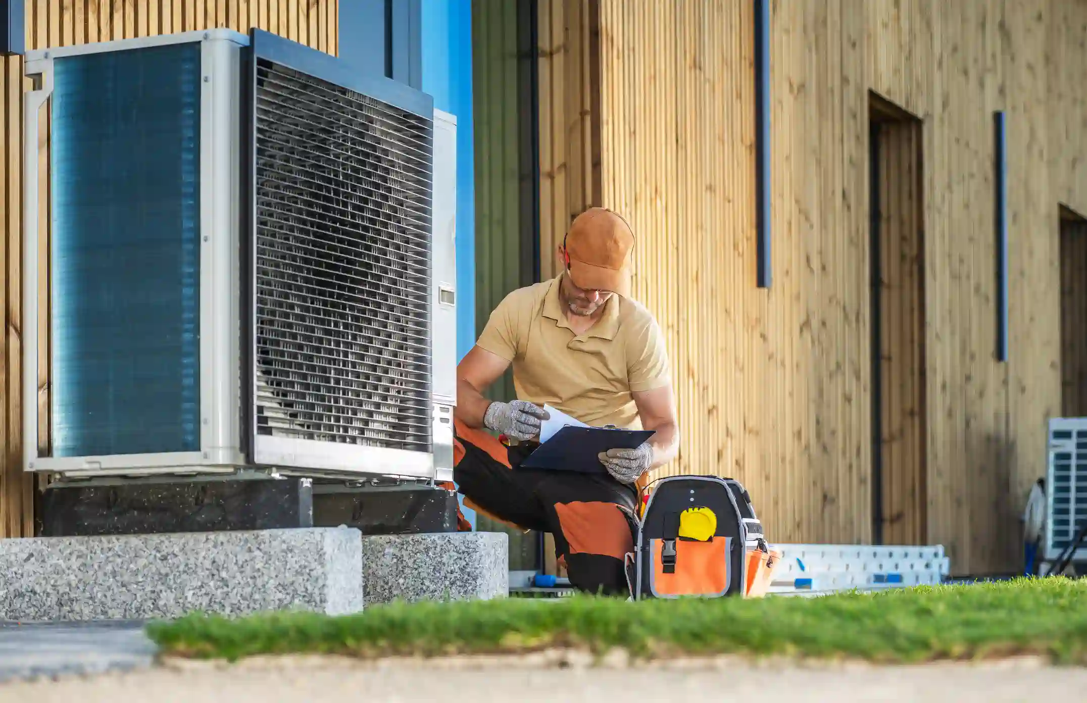 A technician in a tan shirt, orange-and-black work pants, and a baseball cap is crouching by a large outdoor heat pump unit, reviewing paperwork on a clipboard. A tool bag is on the ground next to him, and the building behind has modern wooden siding.