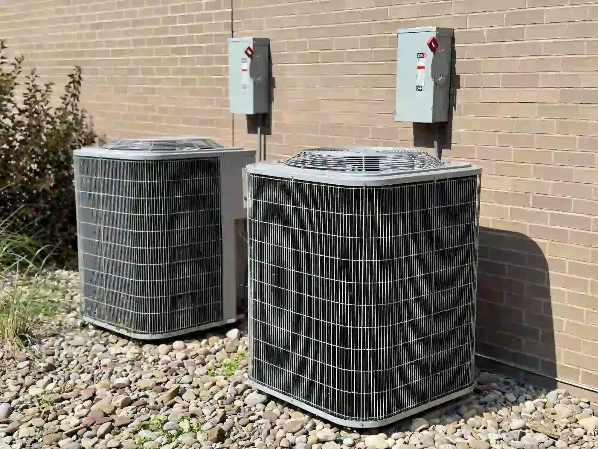 two identical outdoor condensing units (likely for air conditioning or heat pumps) situated next to a brick wall.