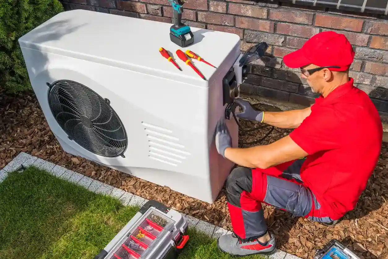 A male technician, dressed in a red shirt, red cap, and gray and red pants, is kneeling next to a large white outdoor heat pump unit. He is wearing gray work gloves and servicing the electrical connections on the side of the unit. A drill and three screwdrivers are resting on top of the heat pump, with a toolbox in the foreground.