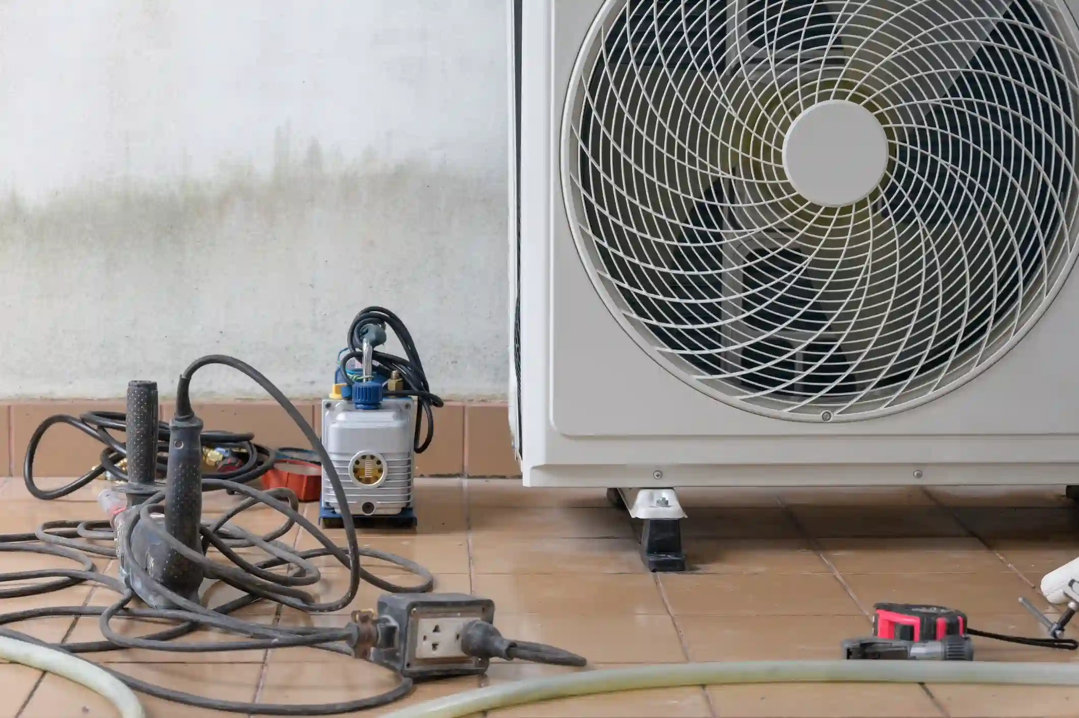 A close-up of an outdoor heat pump installation shows the bottom right side of the white condenser unit, featuring a circular fan grille. On the tiled floor are tools, including a vacuum pump, a tangle of electrical cords, and a power strip, used for the installation.