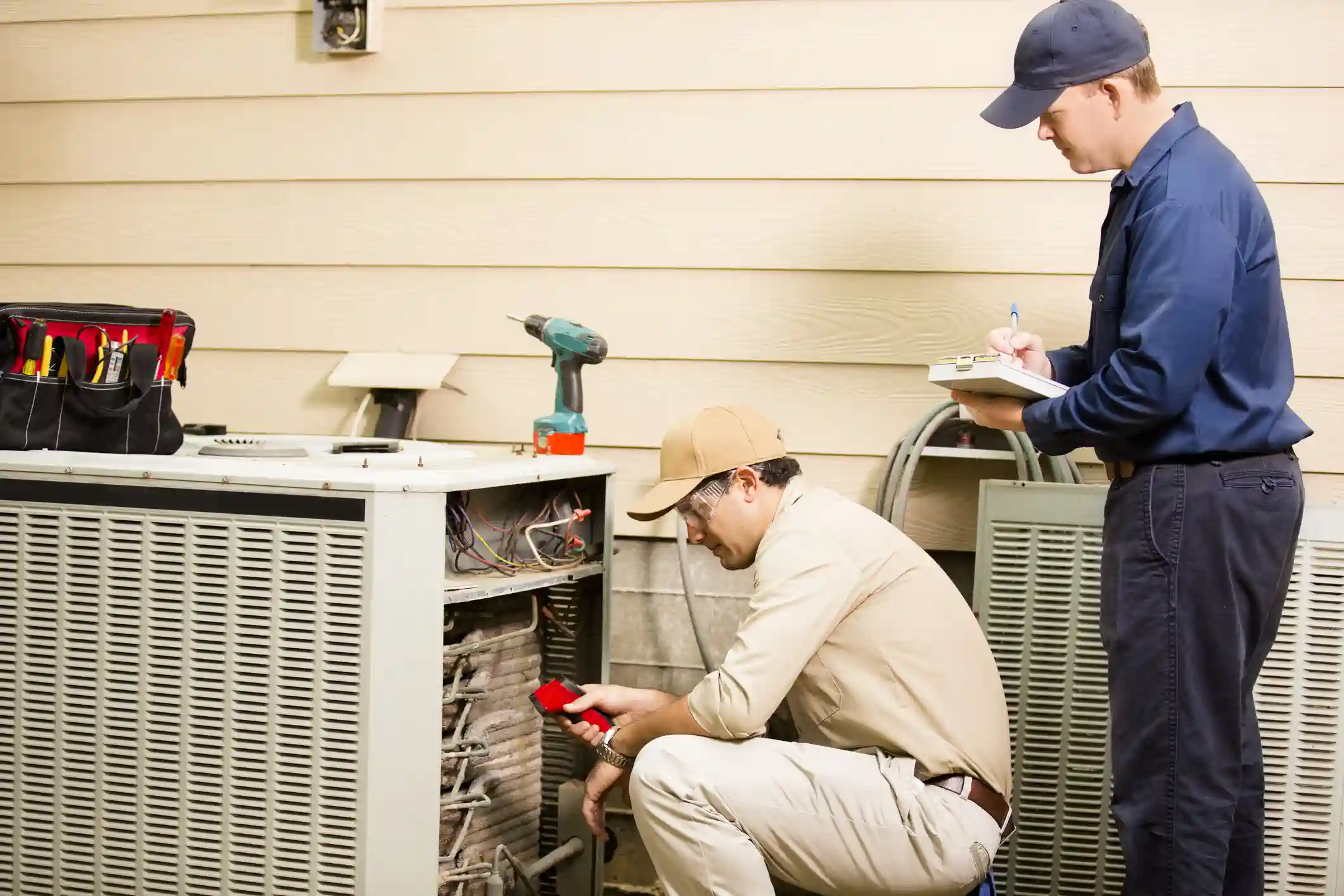 Two HVAC technicians are servicing an outdoor air conditioning condenser unit next to a house with beige siding. One technician is crouching, wearing a tan shirt, cap, and goggles, examining the open unit with a red tool. The second technician stands, wearing a dark blue uniform and cap, taking notes on a clipboard. A tool bag and drill are nearby.