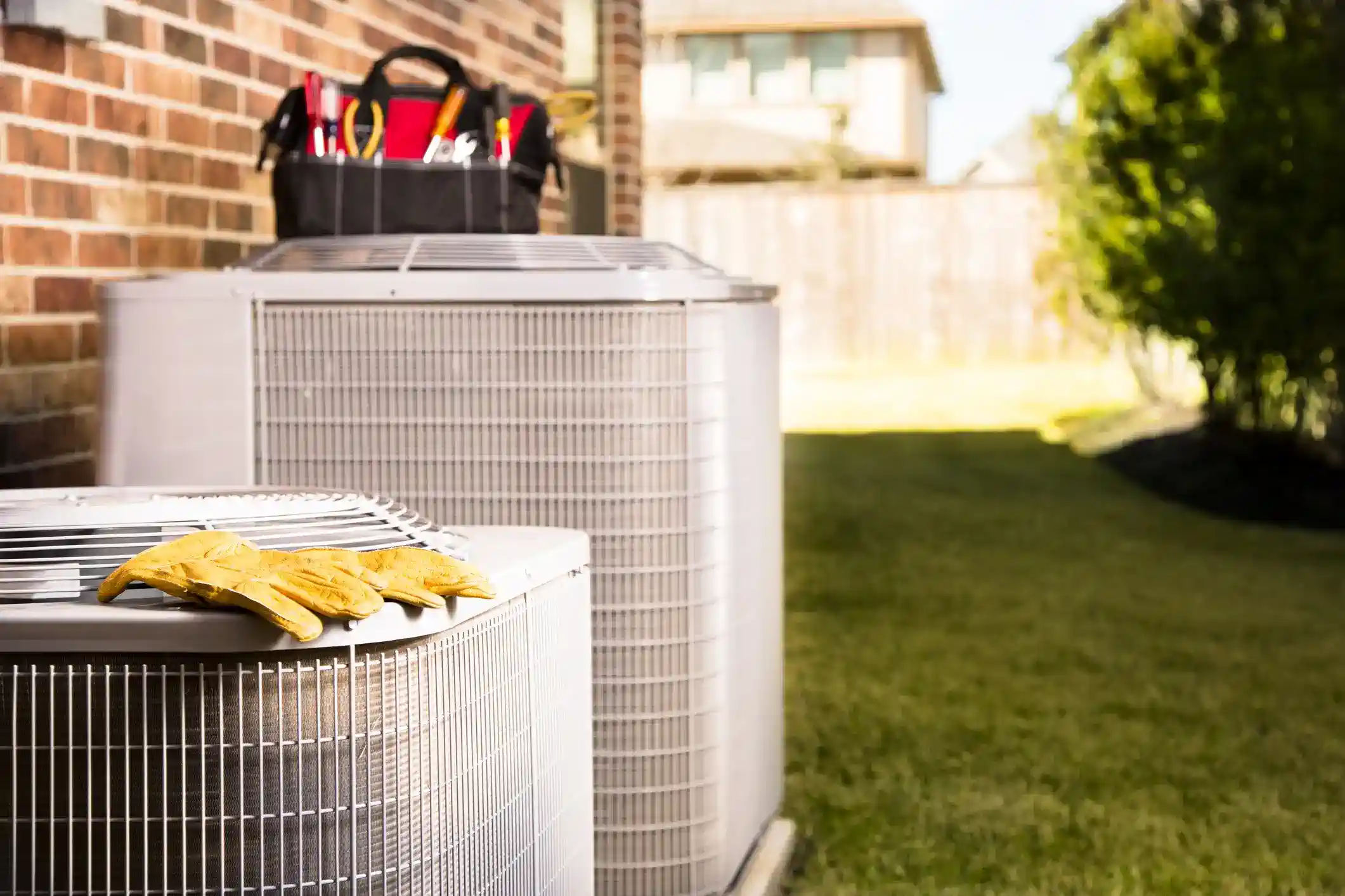 two outdoor condensing units (or heat pumps) placed side-by-side, near a brick wall, against a sunny backdrop of a residential yard.