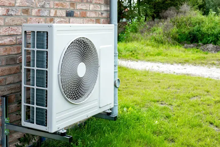 A close-up image showing two outdoor HVAC (Heating, Ventilation, and Air Conditioning) condensing units in a sunny residential yard next to a brick wall. A pair of yellow work gloves is resting on top of the front unit, and a red and black technician's tool bag full of hand tools is sitting on top of the unit further back.