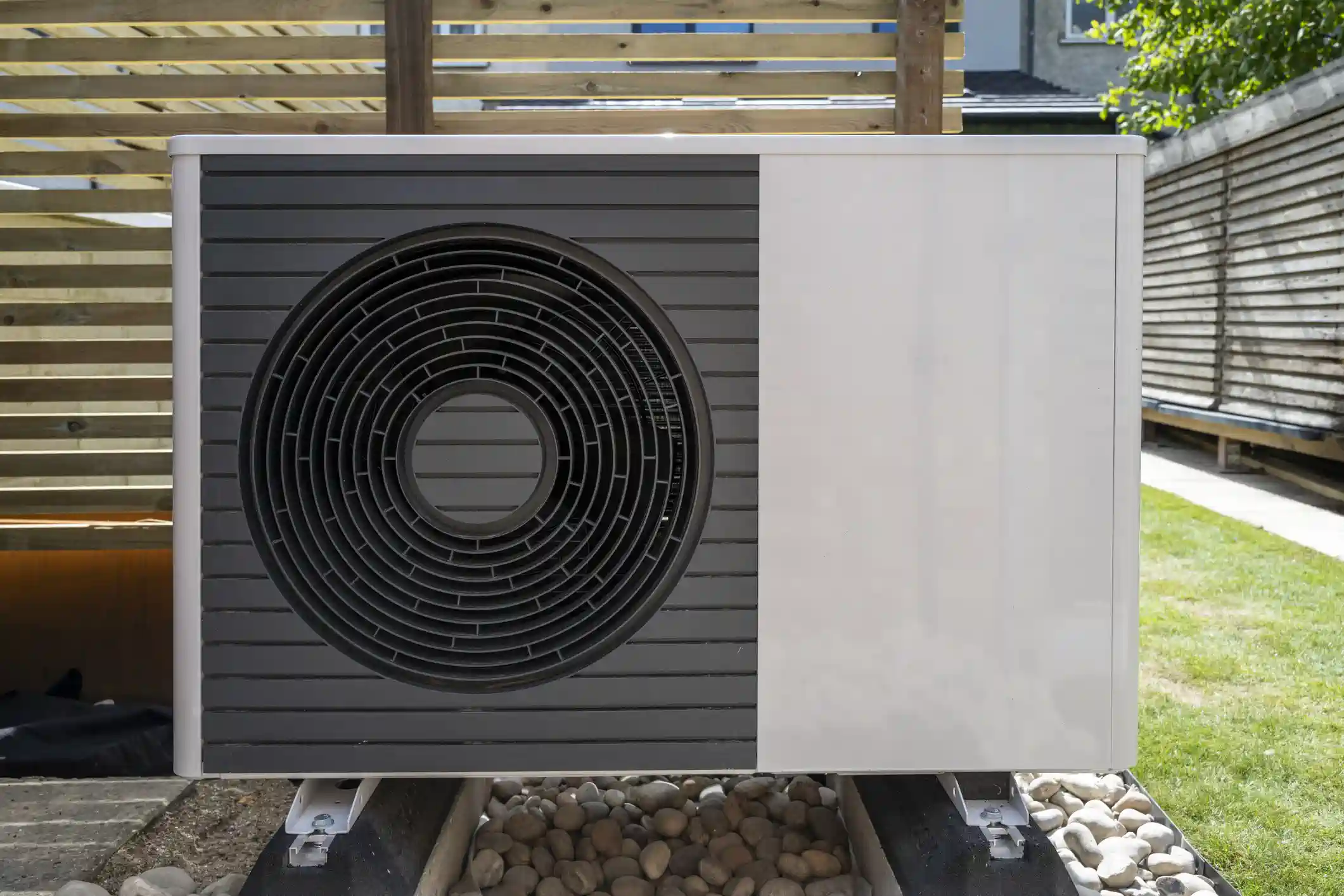 A modern, white and dark grey outdoor unit for an air source heat pump, sitting on metal risers over a bed of light-colored pebbles. The unit has a large circular fan grille on the left, and is placed in a sunny backyard next to a wooden fence or pergola structure.